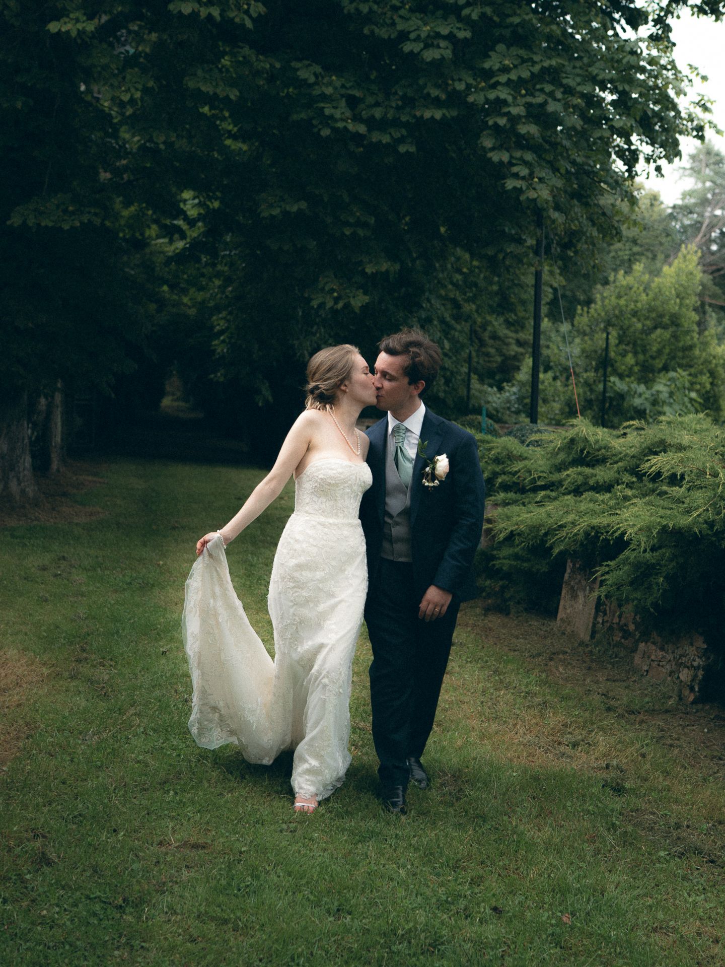 A bride and groom share a romantic kiss while walking through a lush green garden on their wedding day.
