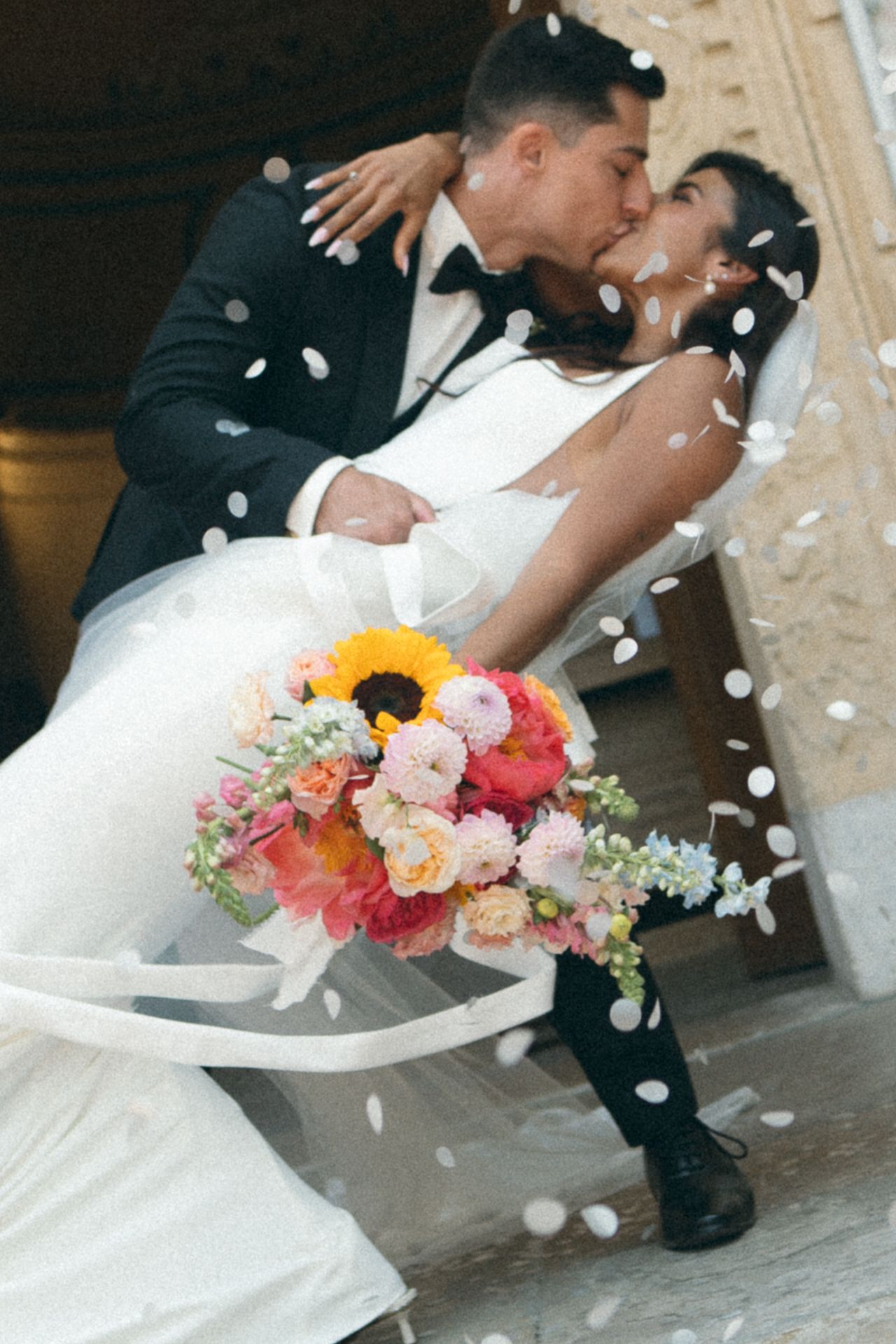 A romantic wedding moment captured with falling petals and vibrant sunflower bouquet during a newlywed couple's kiss.