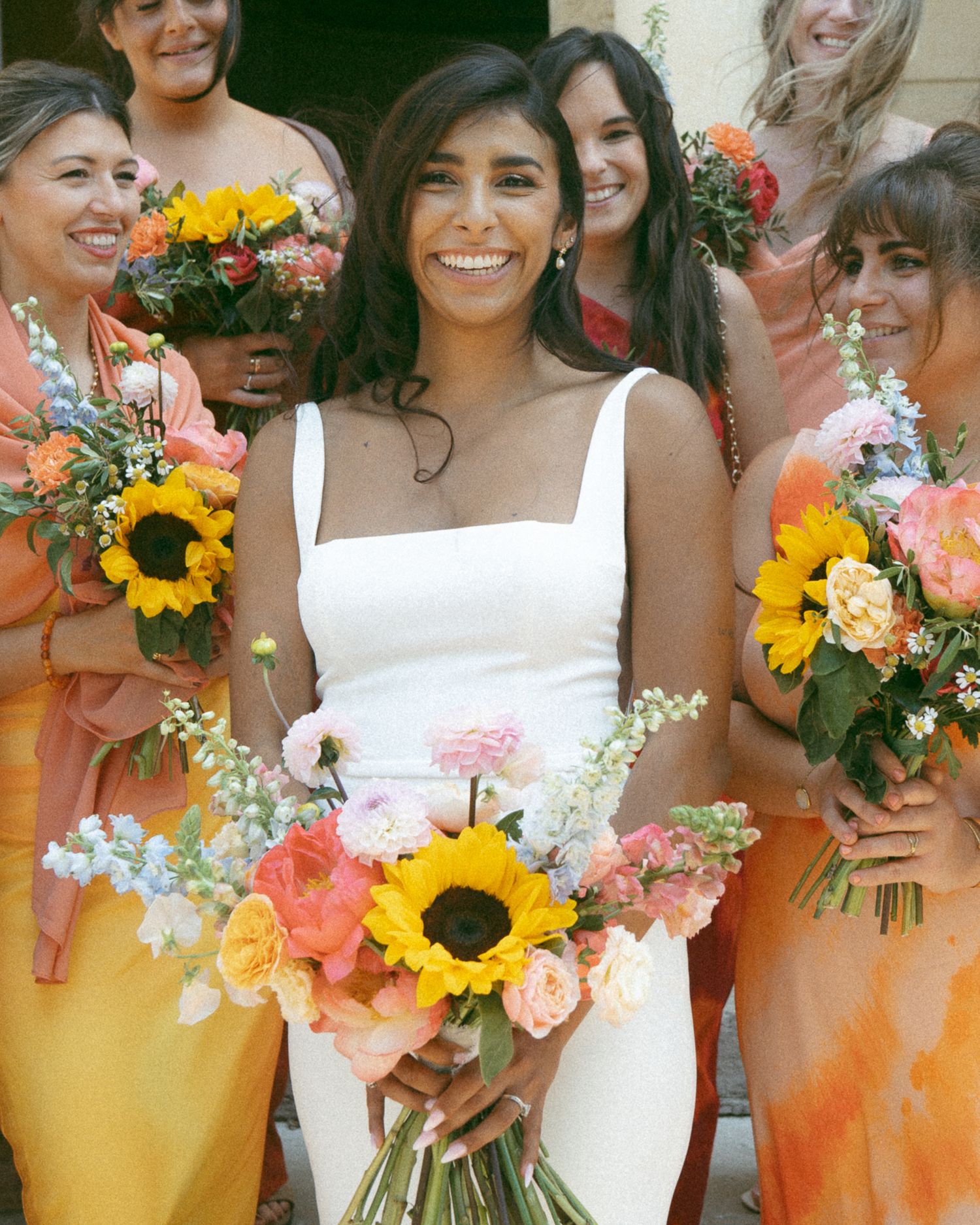 Bridal party in colorful dresses holds vibrant bouquets with sunflowers and mixed flowers against a light background.
