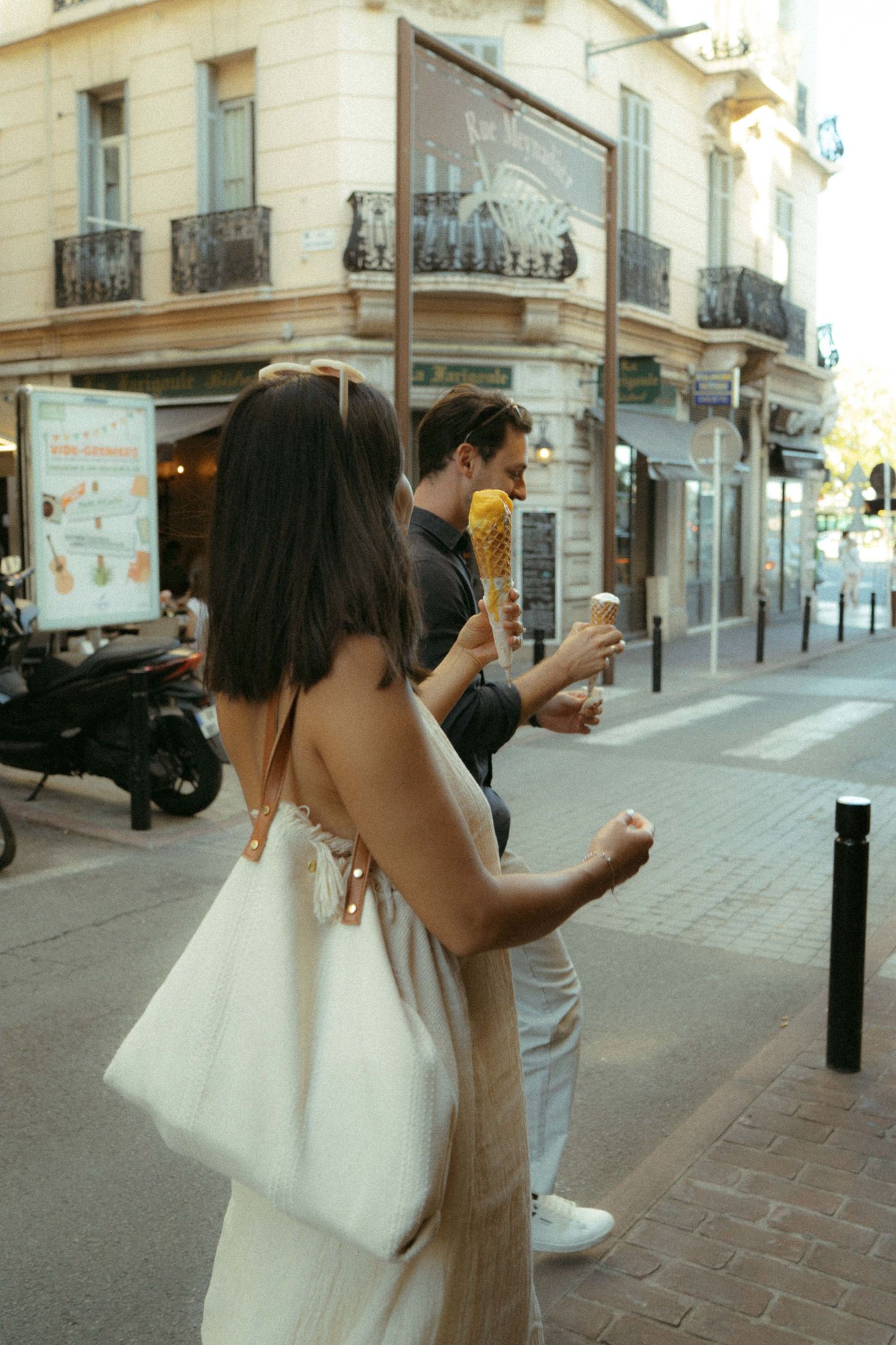 Two people enjoy ice cream cones while walking on a European city street with classic architecture and balconies.