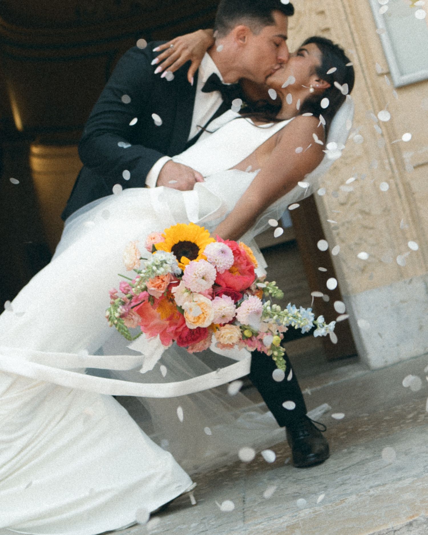 Newlyweds share romantic dip kiss with colorful sunflower bouquet amid falling confetti.