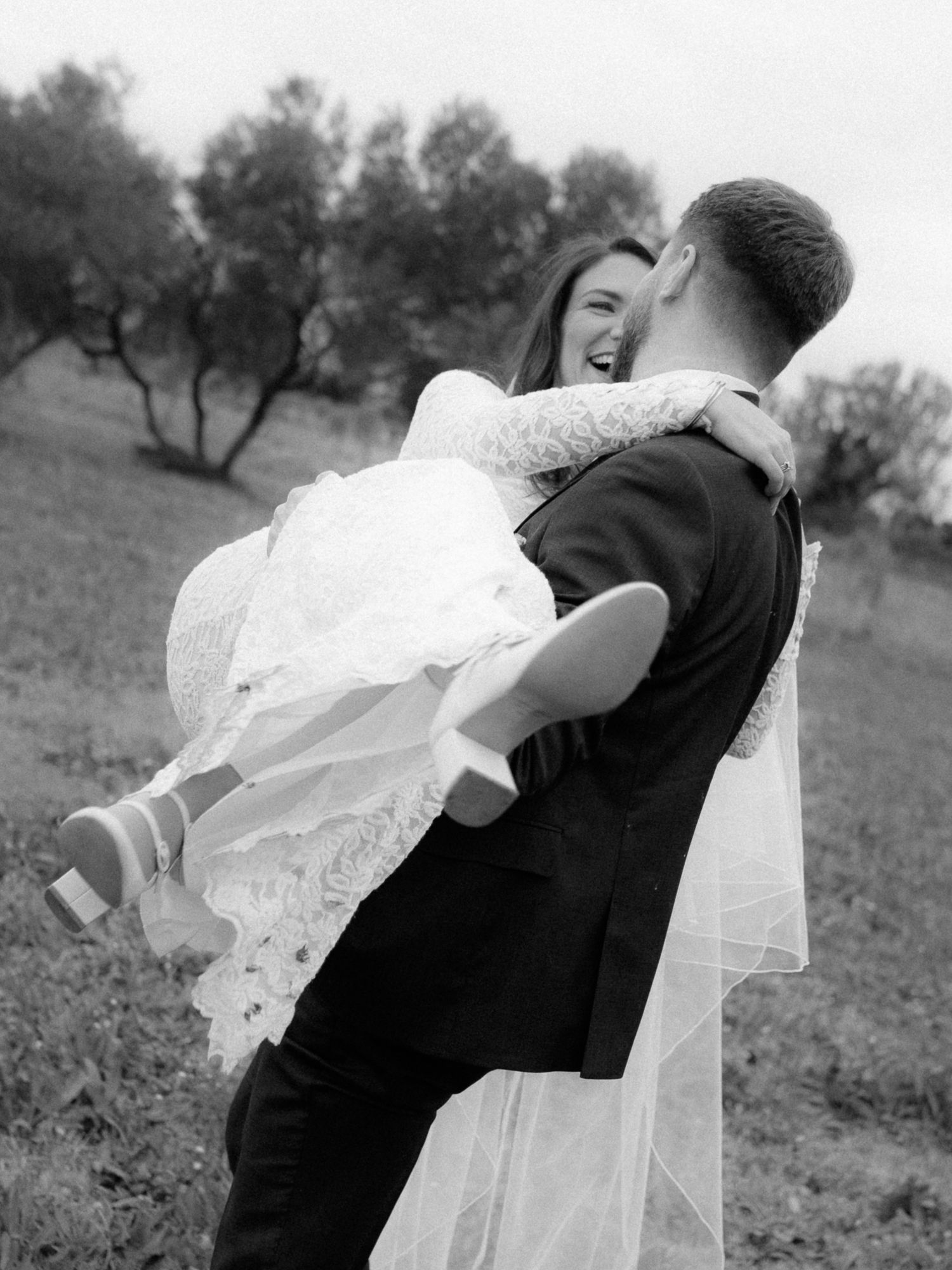 A romantic black and white wedding photo shows a couple embracing joyfully in a natural outdoor setting.
