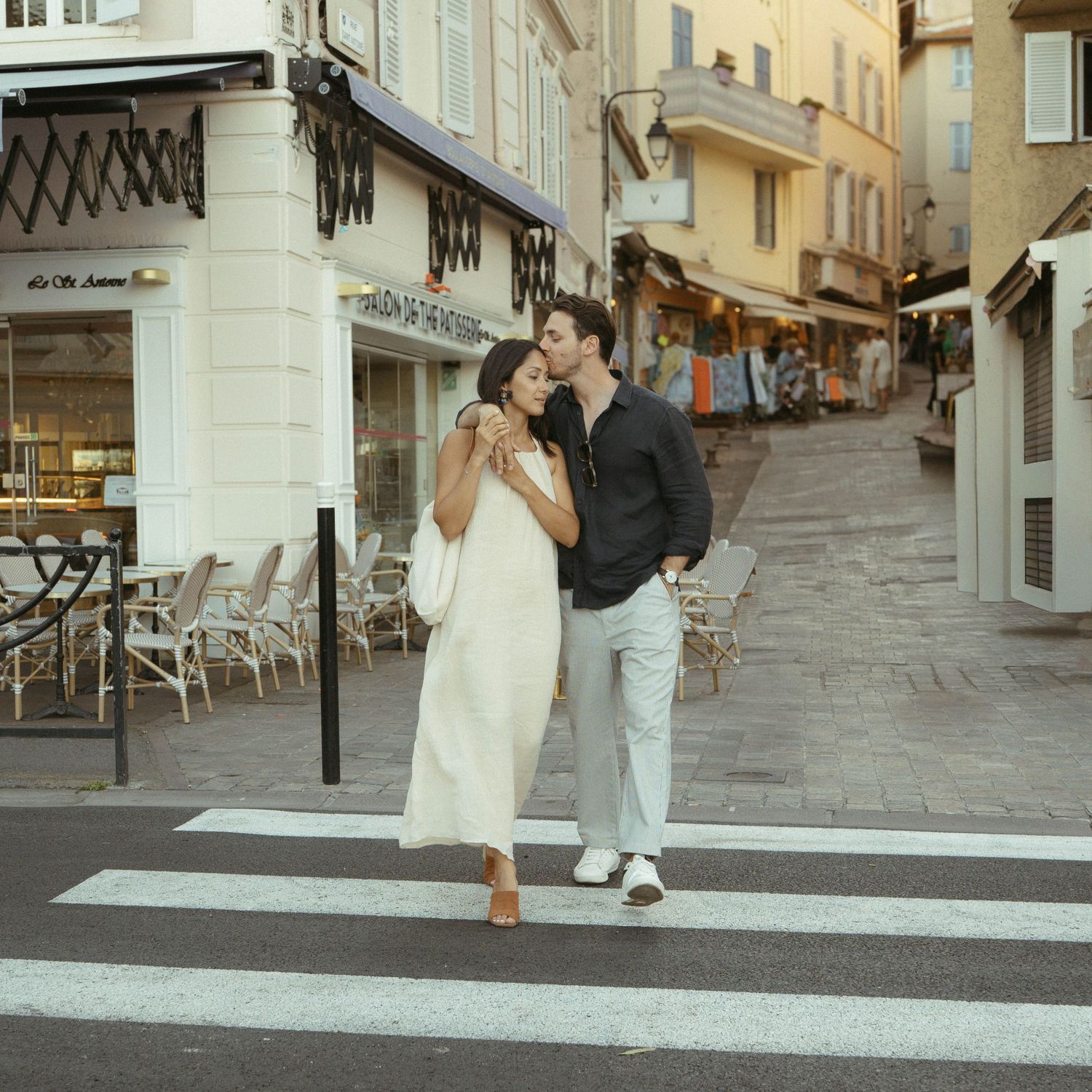 A couple walks down a charming European cobblestone street lined with cafes and shops on a sunny day.