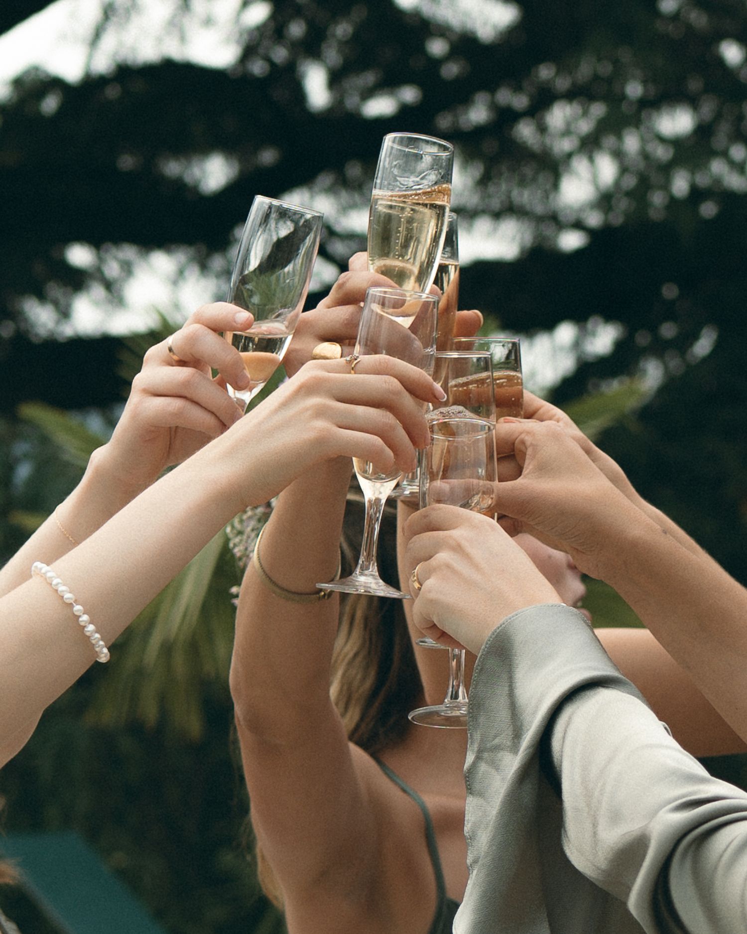 Multiple hands raise champagne glasses in a celebratory toast against a blurred outdoor background.