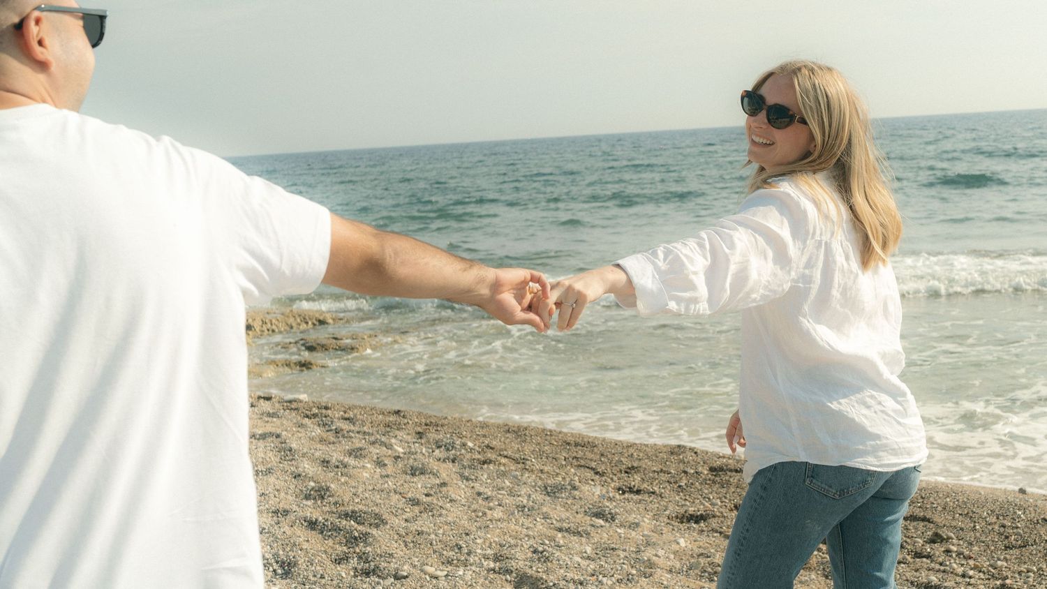 Person in white shirt and jeans walks along sandy beach with ocean waves in the background.
