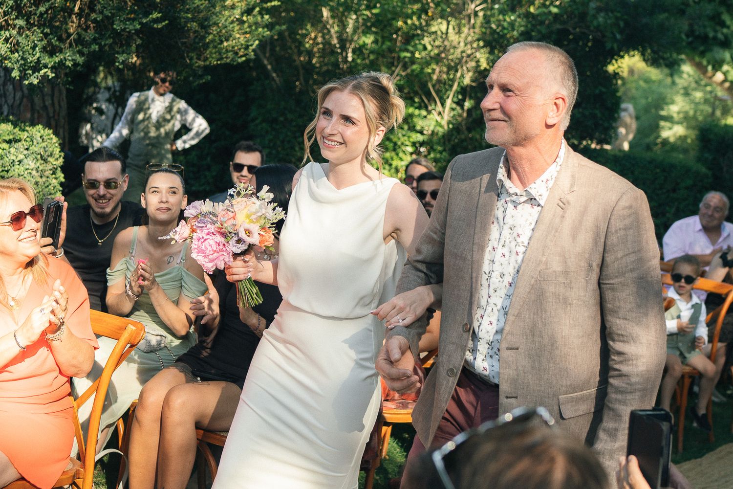 Bride in white dress holding pink bouquet walks with groom in tan suit at outdoor wedding ceremony.