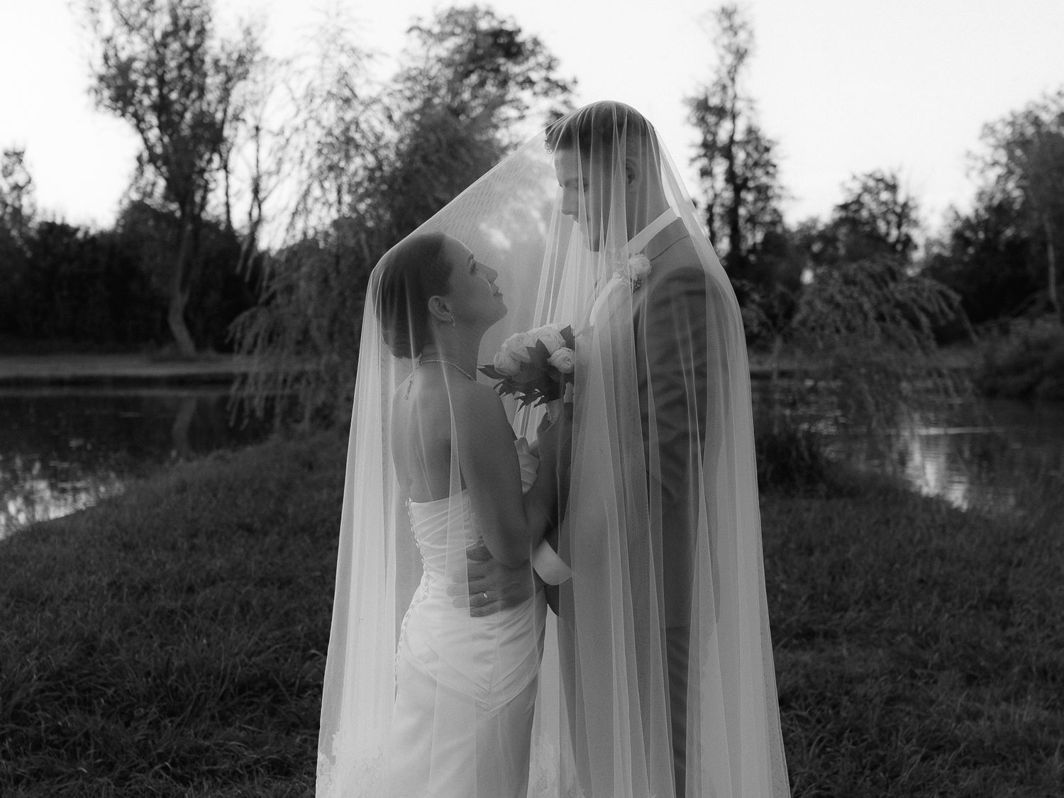 Black and white photo of two brides sharing a kiss under a flowing veil outdoors near a pond.