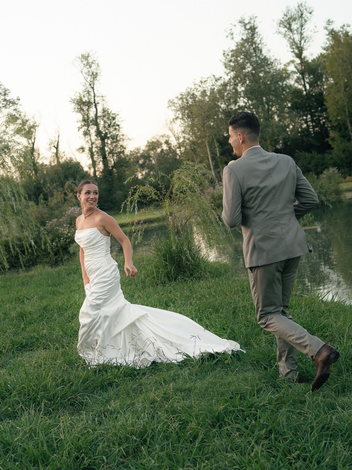 Bride in white gown and groom in tan suit run toward each other on green grass near a pond at golden hour.