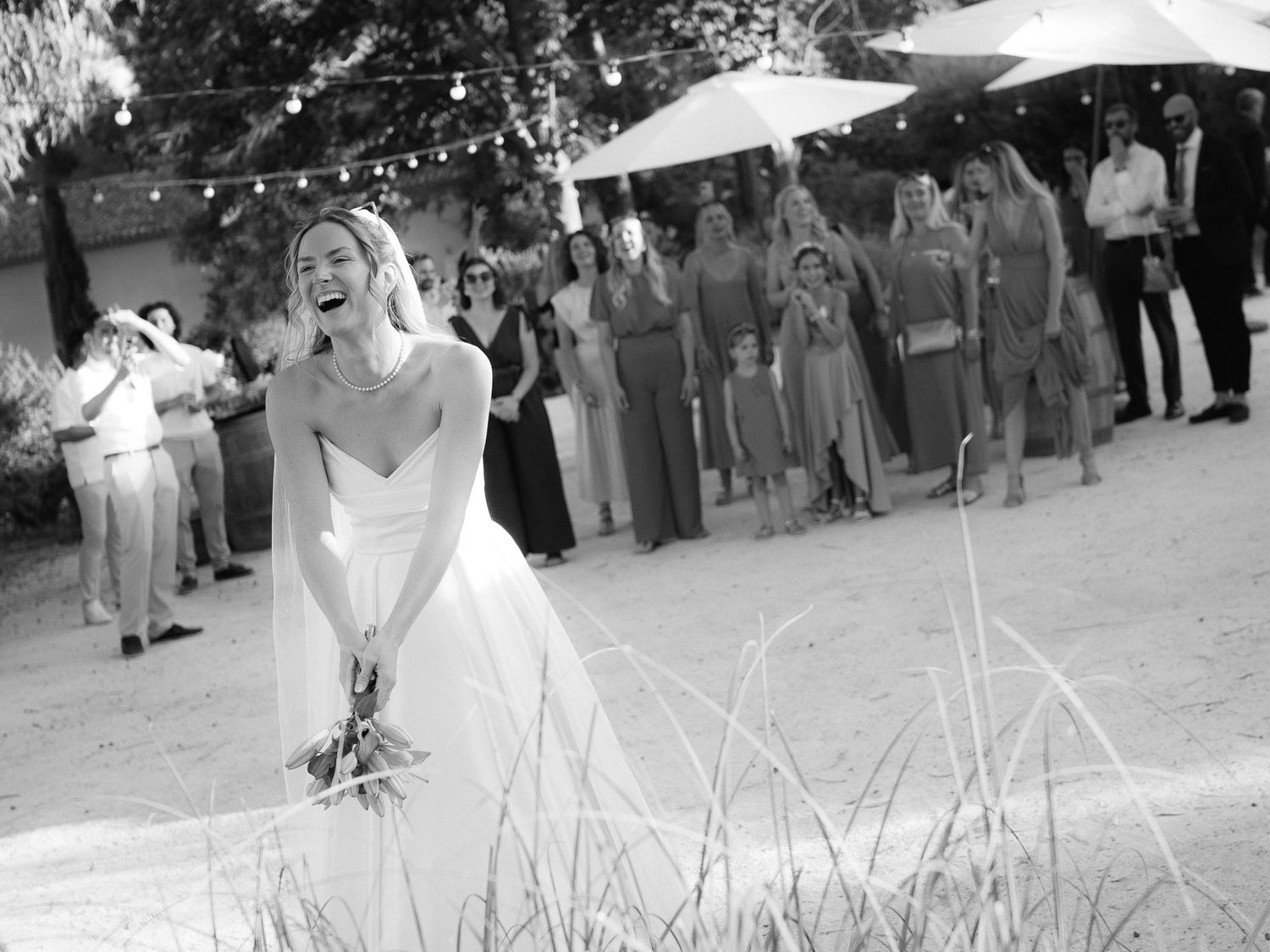 Black and white photo of a bride laughing while tossing her bouquet at an outdoor wedding reception.