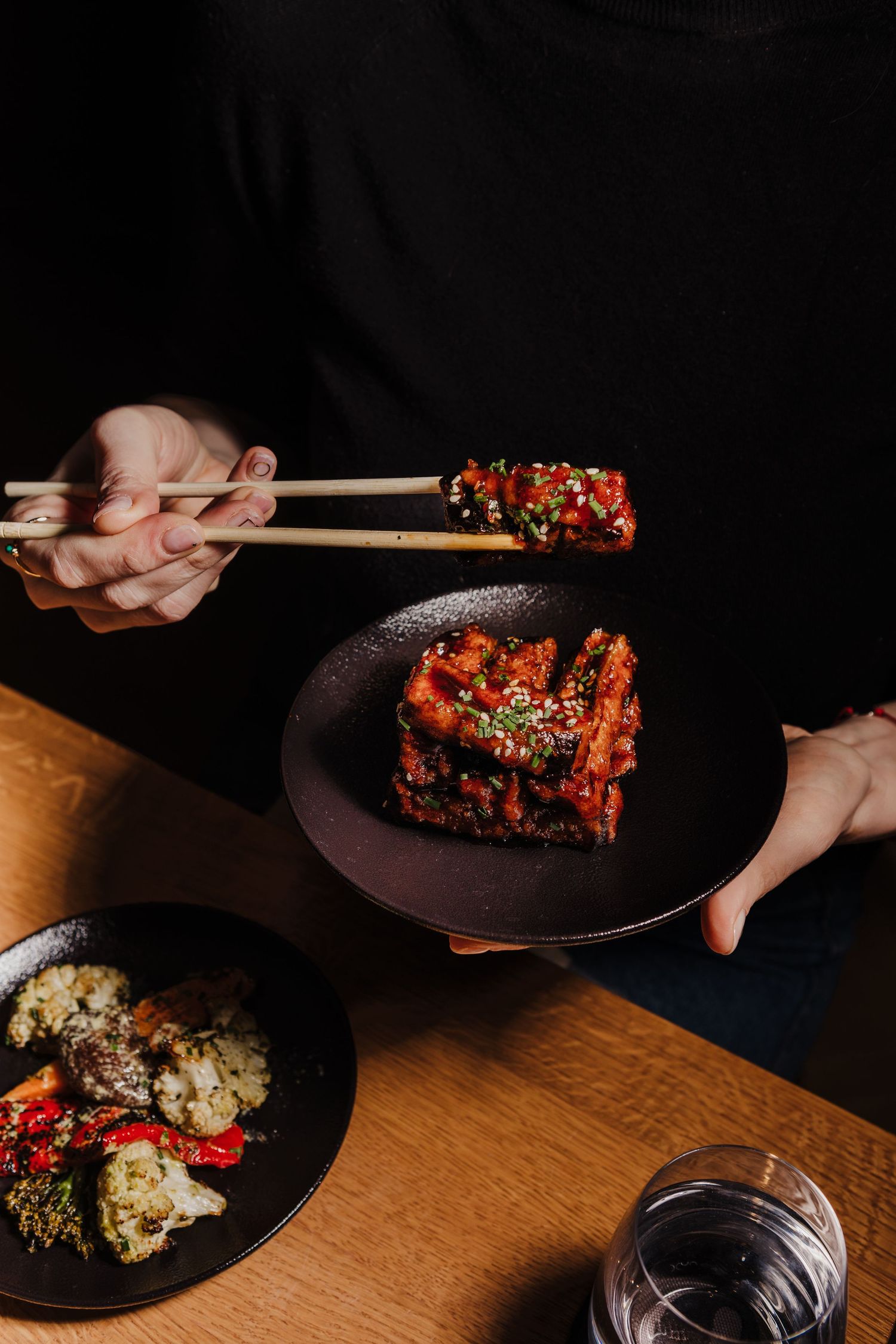 Chopsticks lifting glazed meat dish from dark plate with wine glass on wooden table.