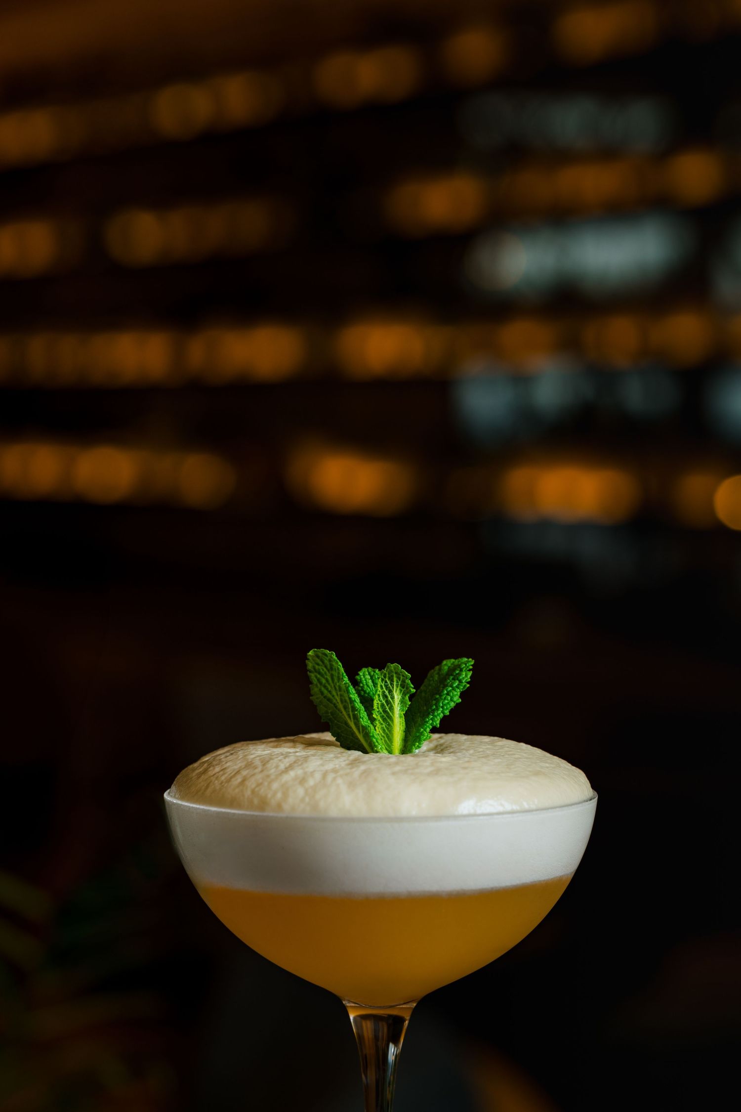 Yellow cocktail with foam head and mint garnish in coupe glass against dark background.