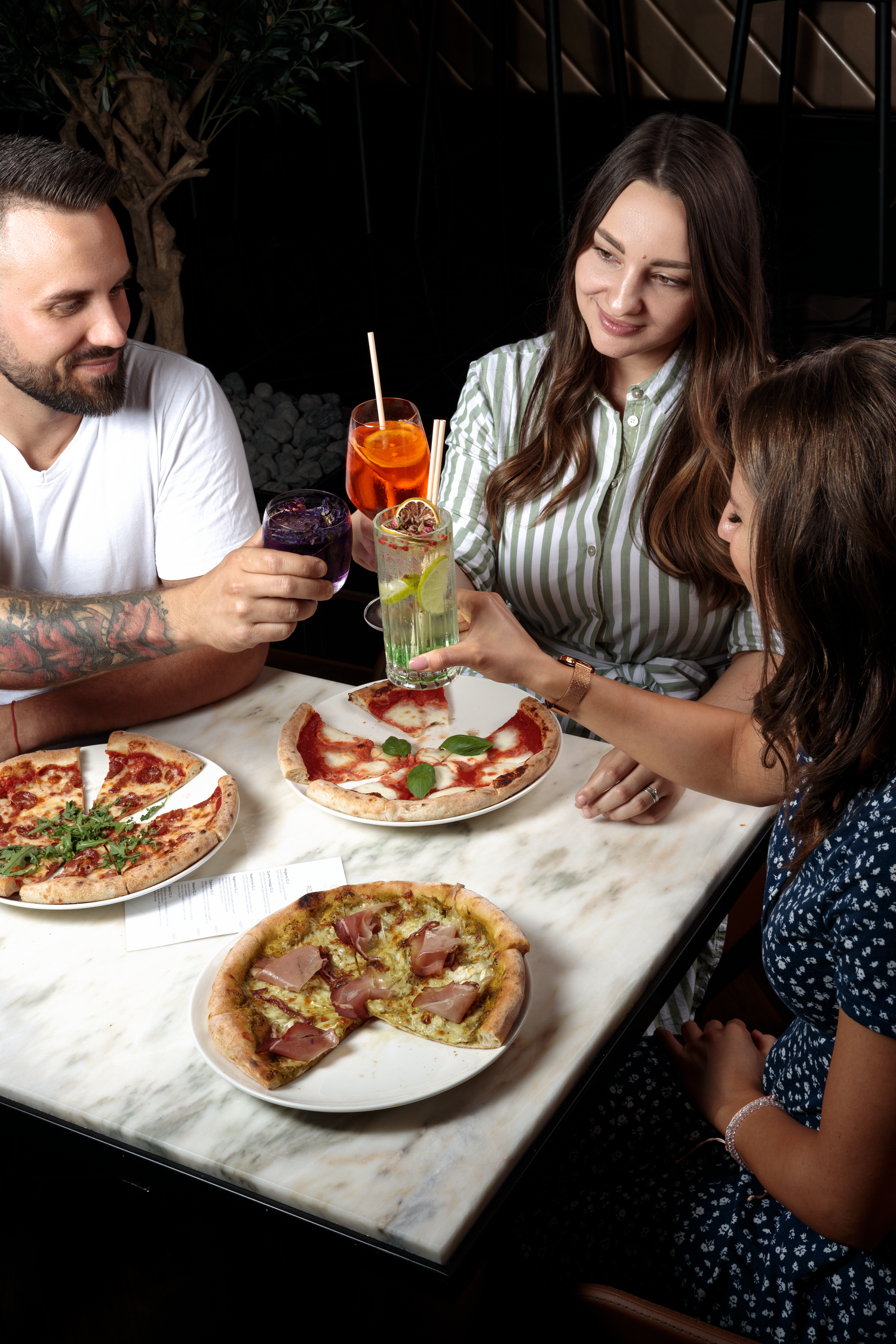 Family enjoying fresh Italian pizzas at a restaurant with colorful cocktails on a marble table.
