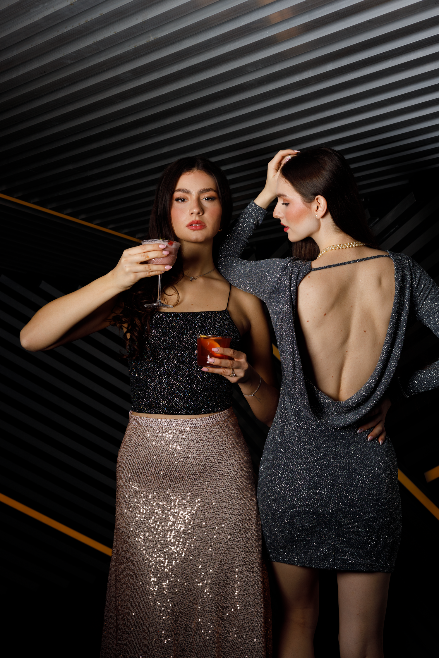 Two women in elegant evening attire pose with cocktails against a dark striped background at a nightclub.