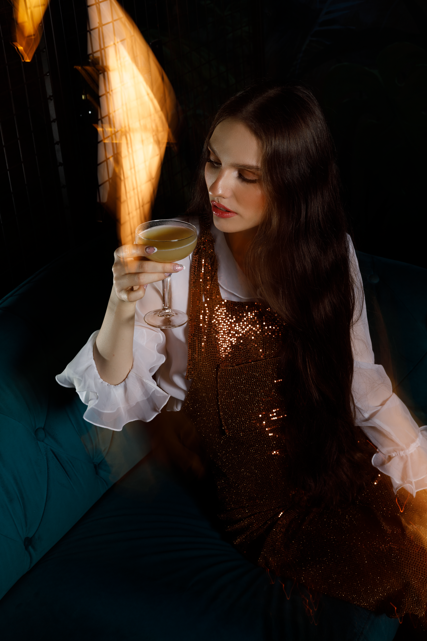 A person in a sequined dress holds a cocktail glass while sitting in moody lighting with dramatic shadows and golden glow.