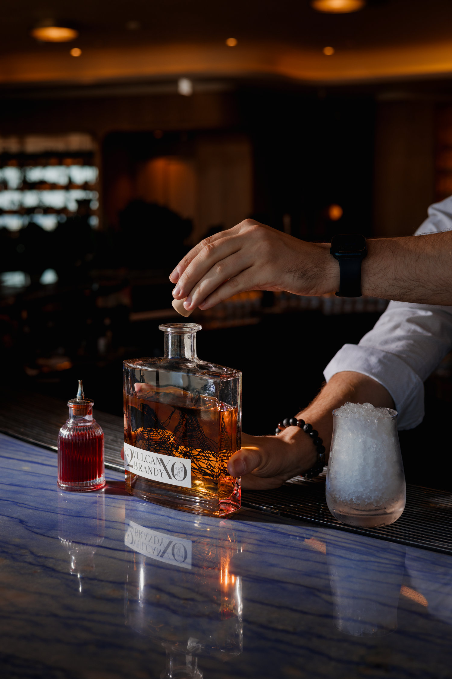 A bartender opens a crystal decanter of amber liquor on a dark marble bar counter with a small red bottle nearby.
