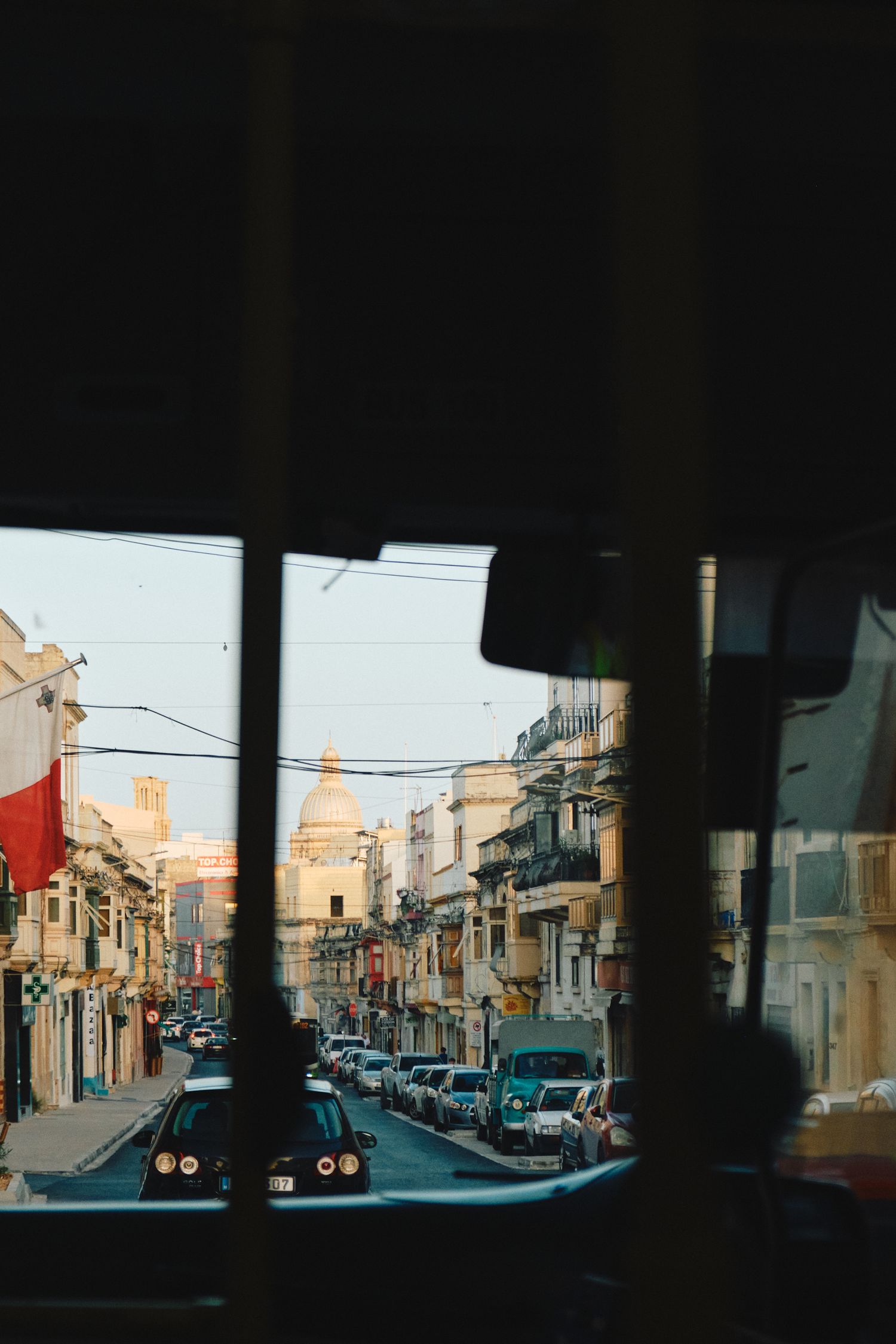 Street view through car windshield of a Mediterranean city with parked cars and buildings.
