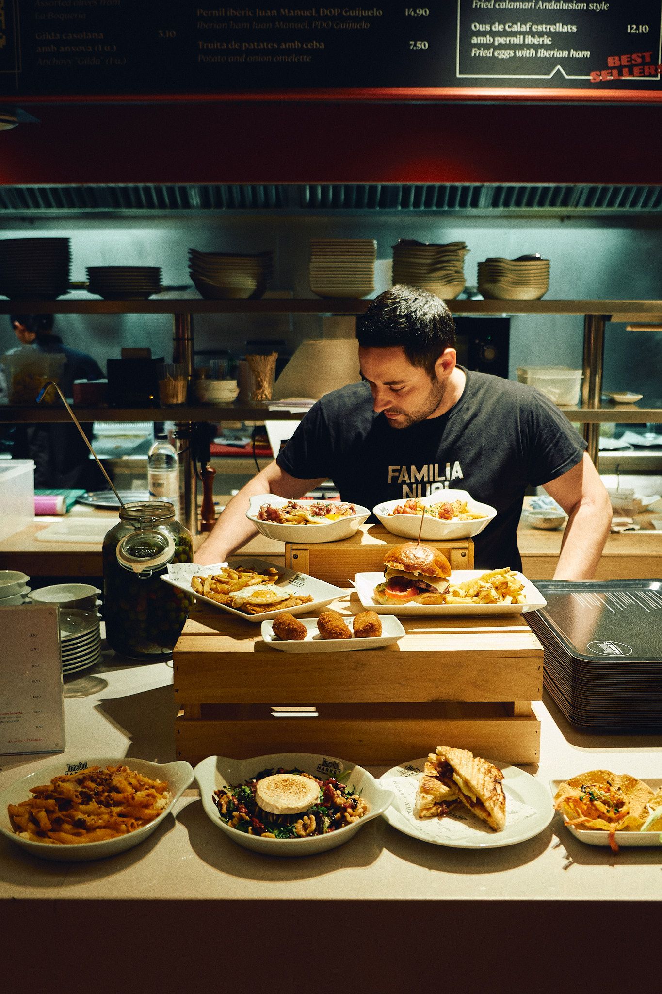 Chef arranging multiple Spanish tapas dishes on a wooden counter in a busy restaurant kitchen.
