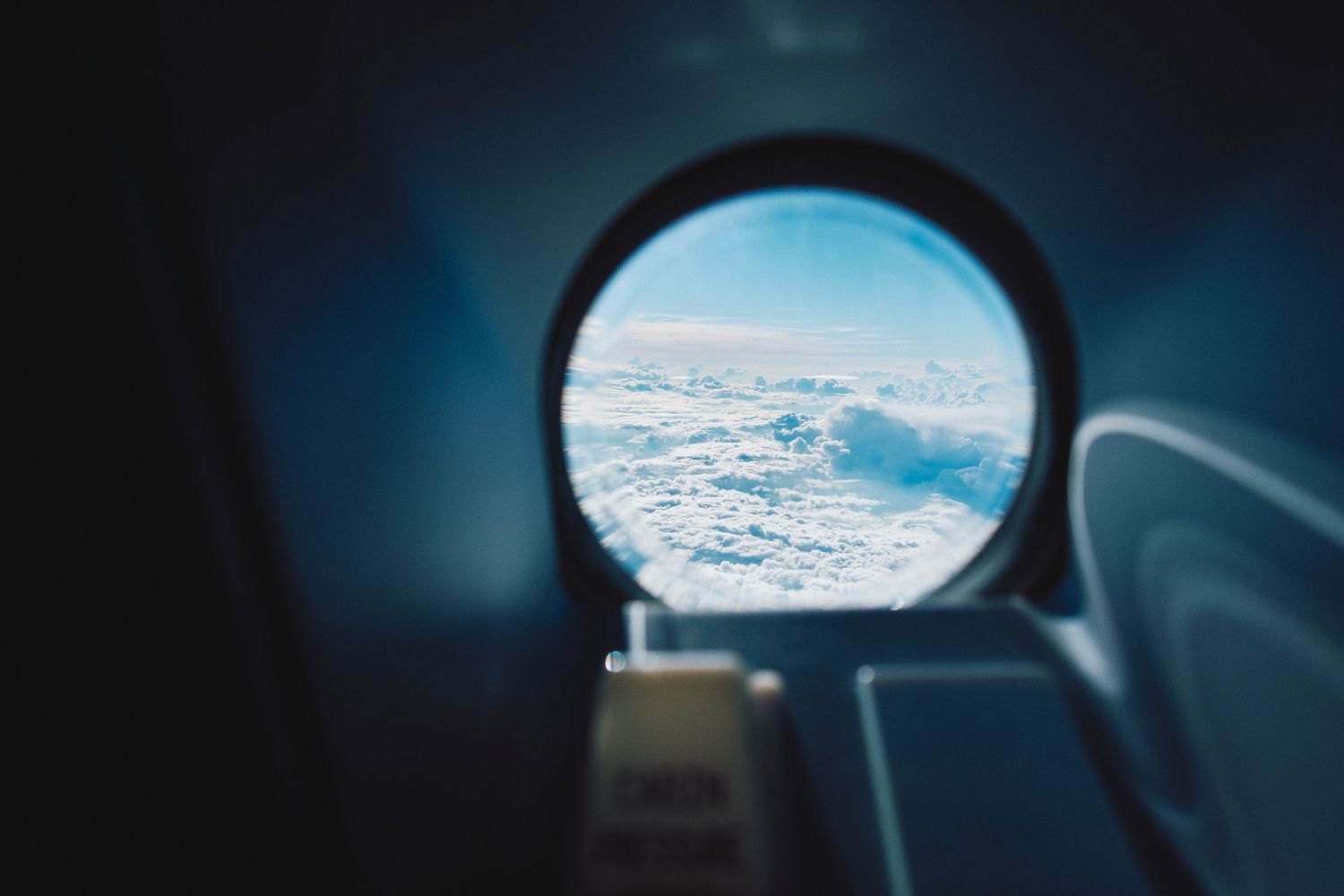 View of fluffy white clouds through an airplane window porthole against a blue sky.