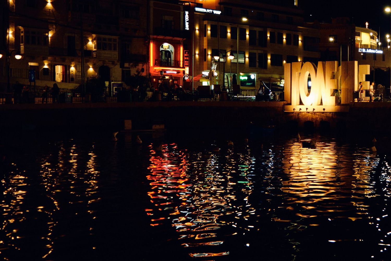 Nighttime reflection of city lights on dark water with illuminated buildings along waterfront.