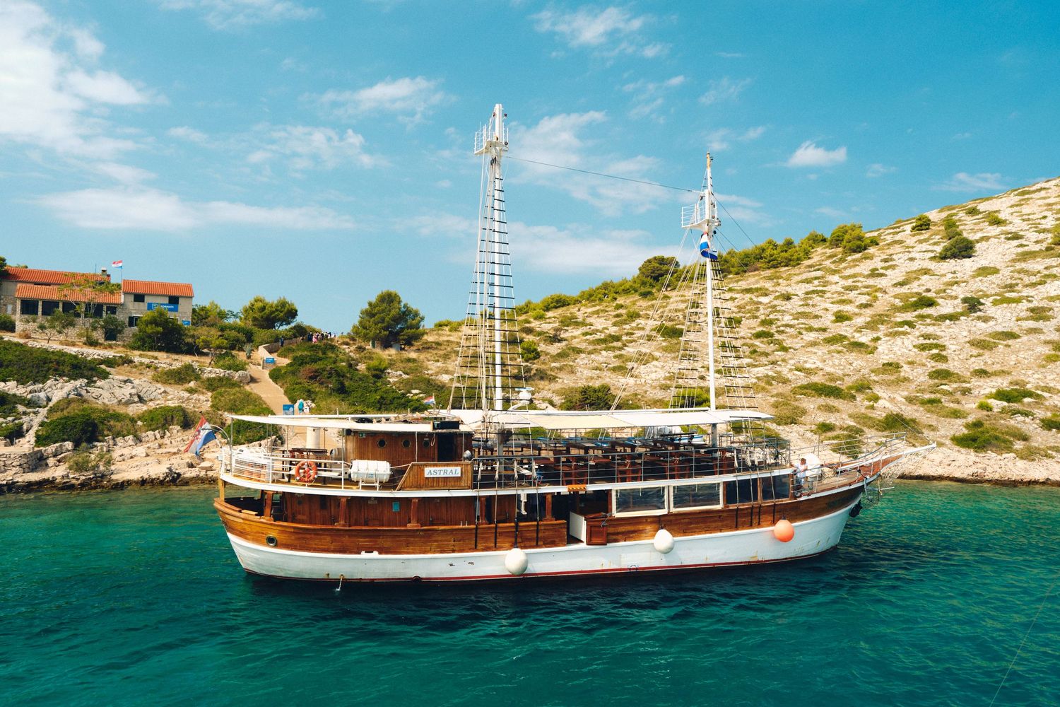 Traditional wooden cruise ship anchored in turquoise Mediterranean waters near rocky coastline.