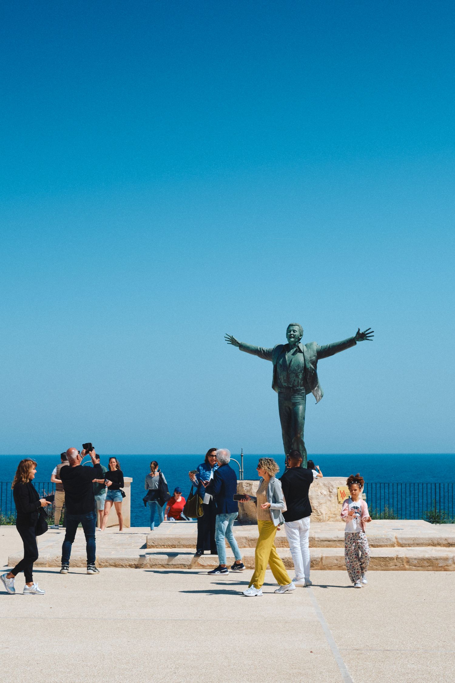 Bronze statue with outstretched arms on seaside promenade with tourists gathering around.