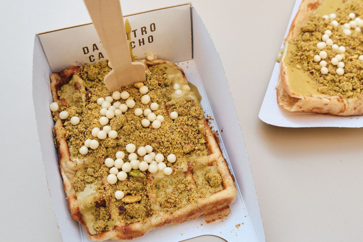 Artisanal toast topped with pistachio spread and white chocolate pearls in a takeout box.