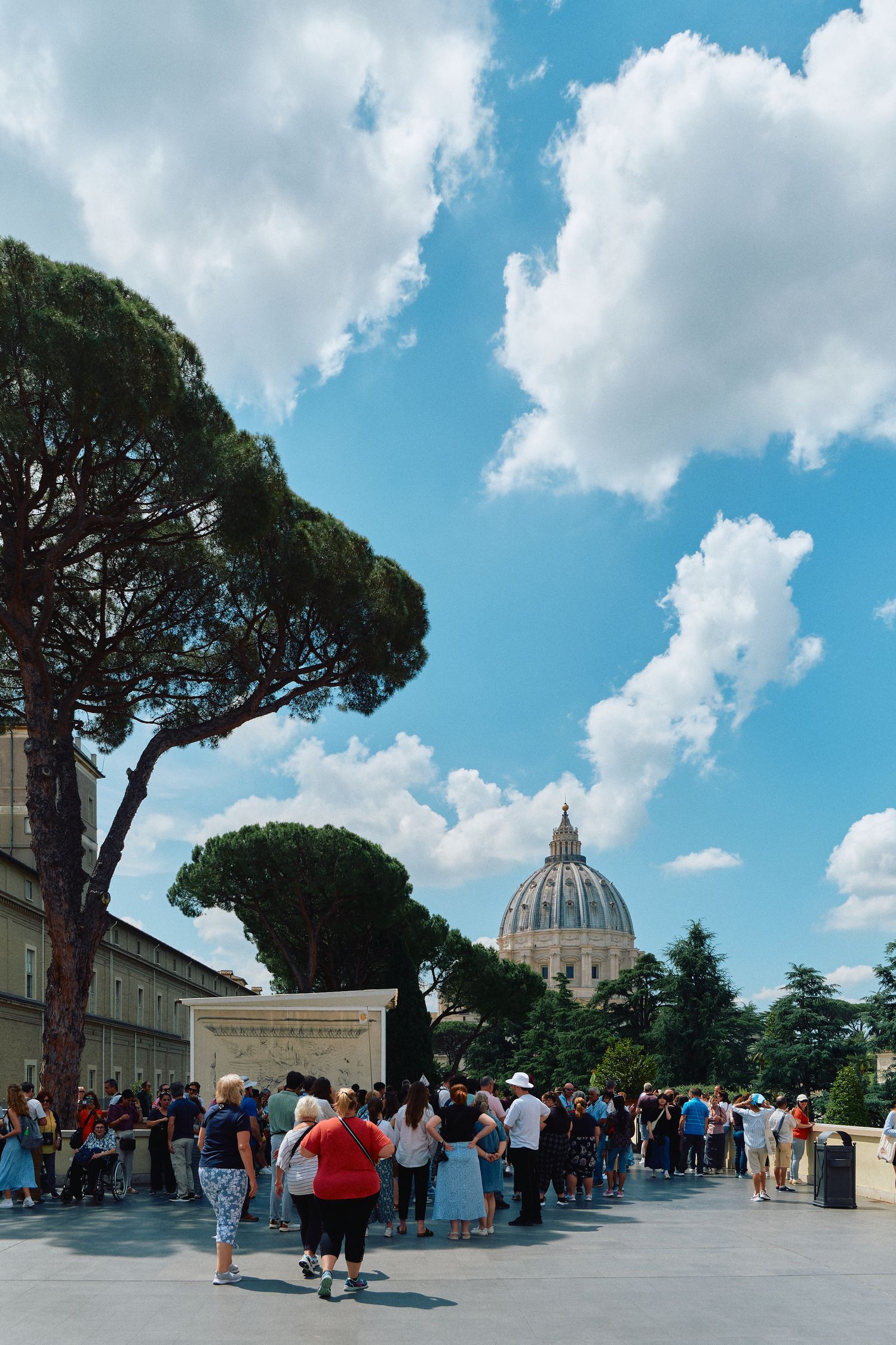 Crowds of tourists gathering outside St. Peter's Basilica dome in Vatican City on a sunny day.