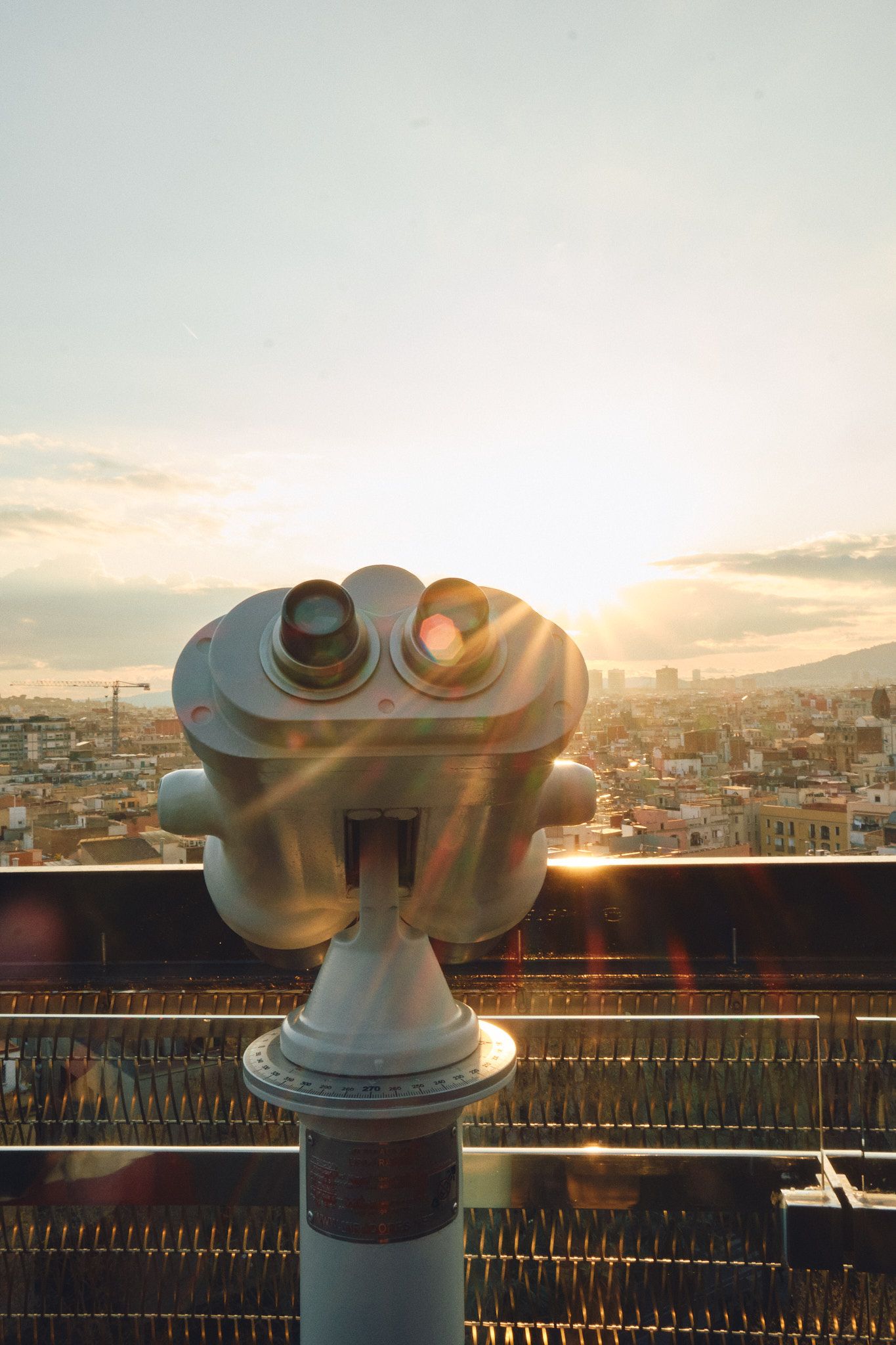 Coin-operated binoculars on a rooftop overlooking a city skyline at golden sunset with warm lens flare.