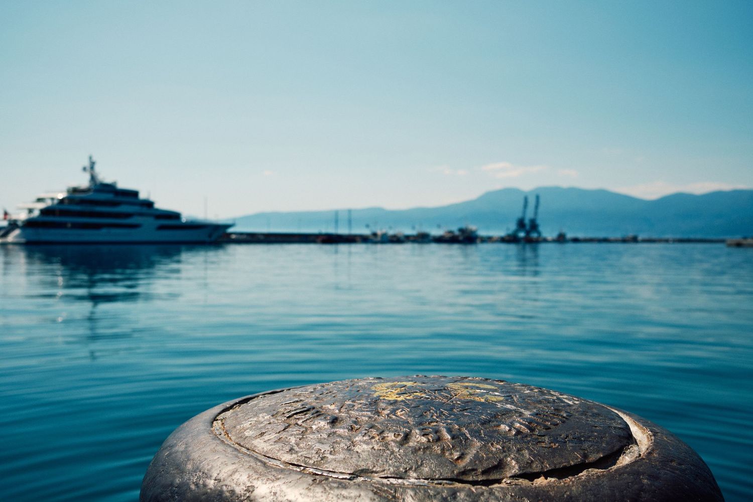 Luxury yacht anchored in calm blue harbor waters with mountains in distance.