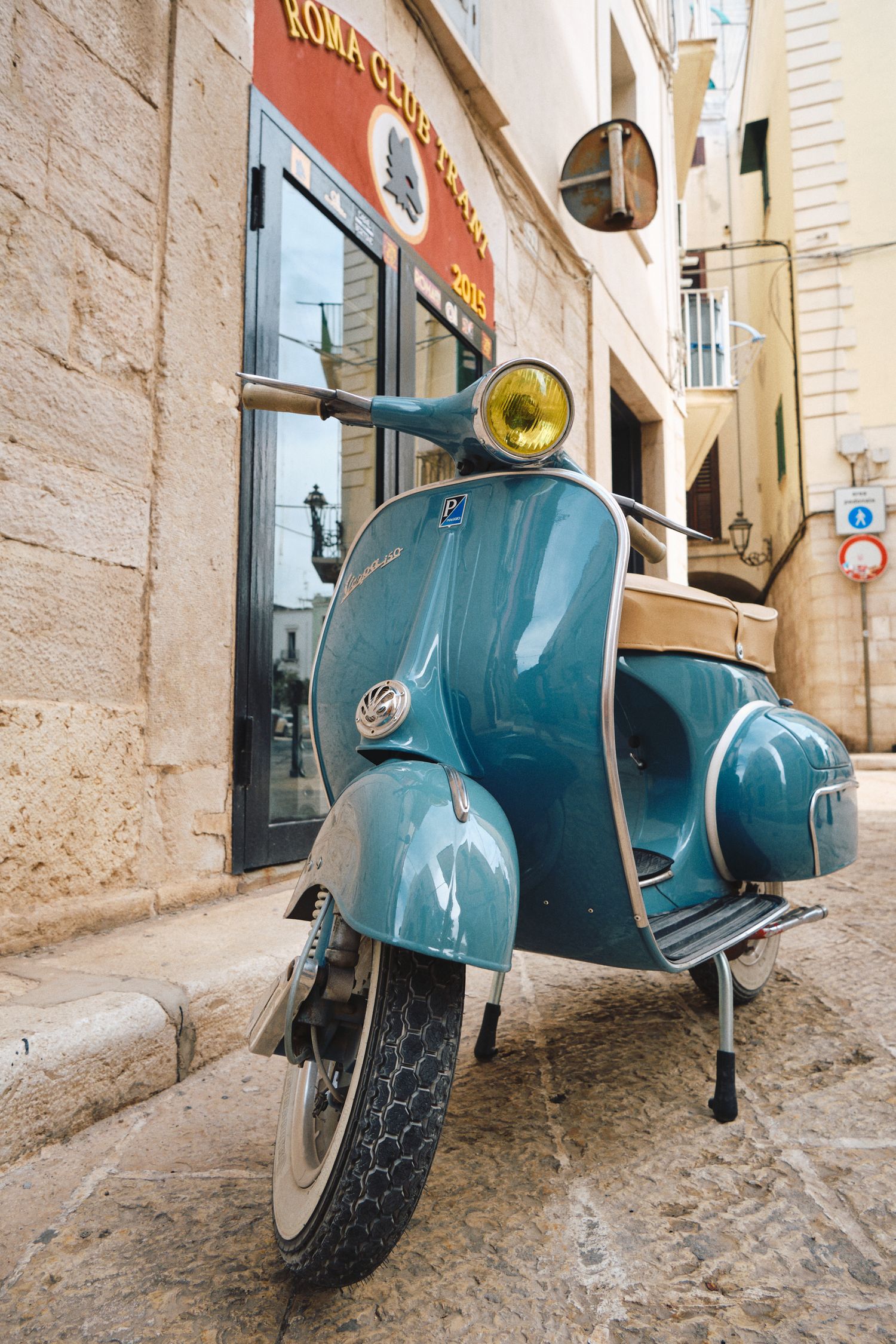 A classic turquoise Vespa scooter parked in front of a Mediterranean building facade.