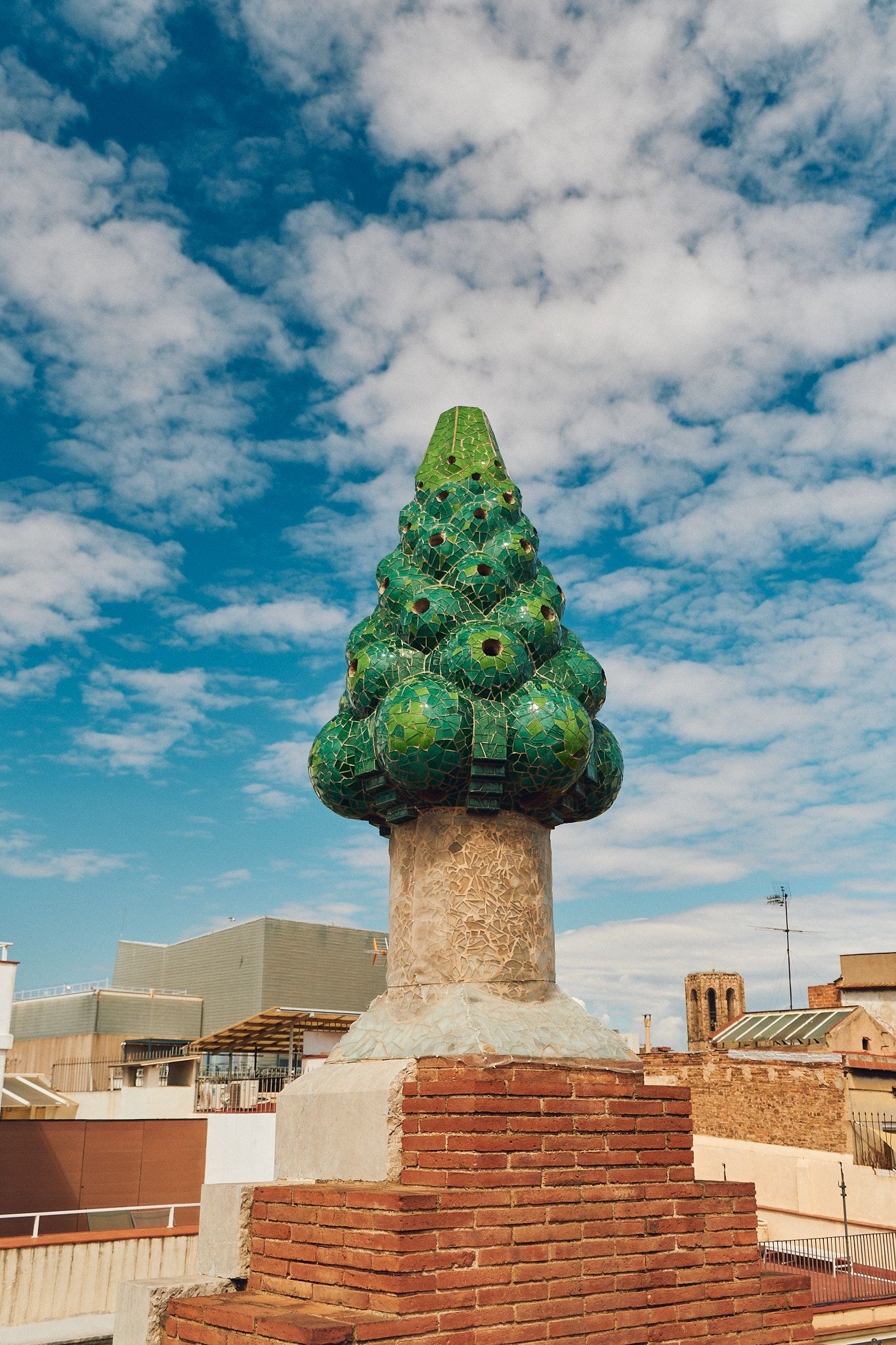 Green mosaic pine cone chimney sculpture on brick base against blue sky, likely Palau Guell rooftop by Gaudi, Barcelona.