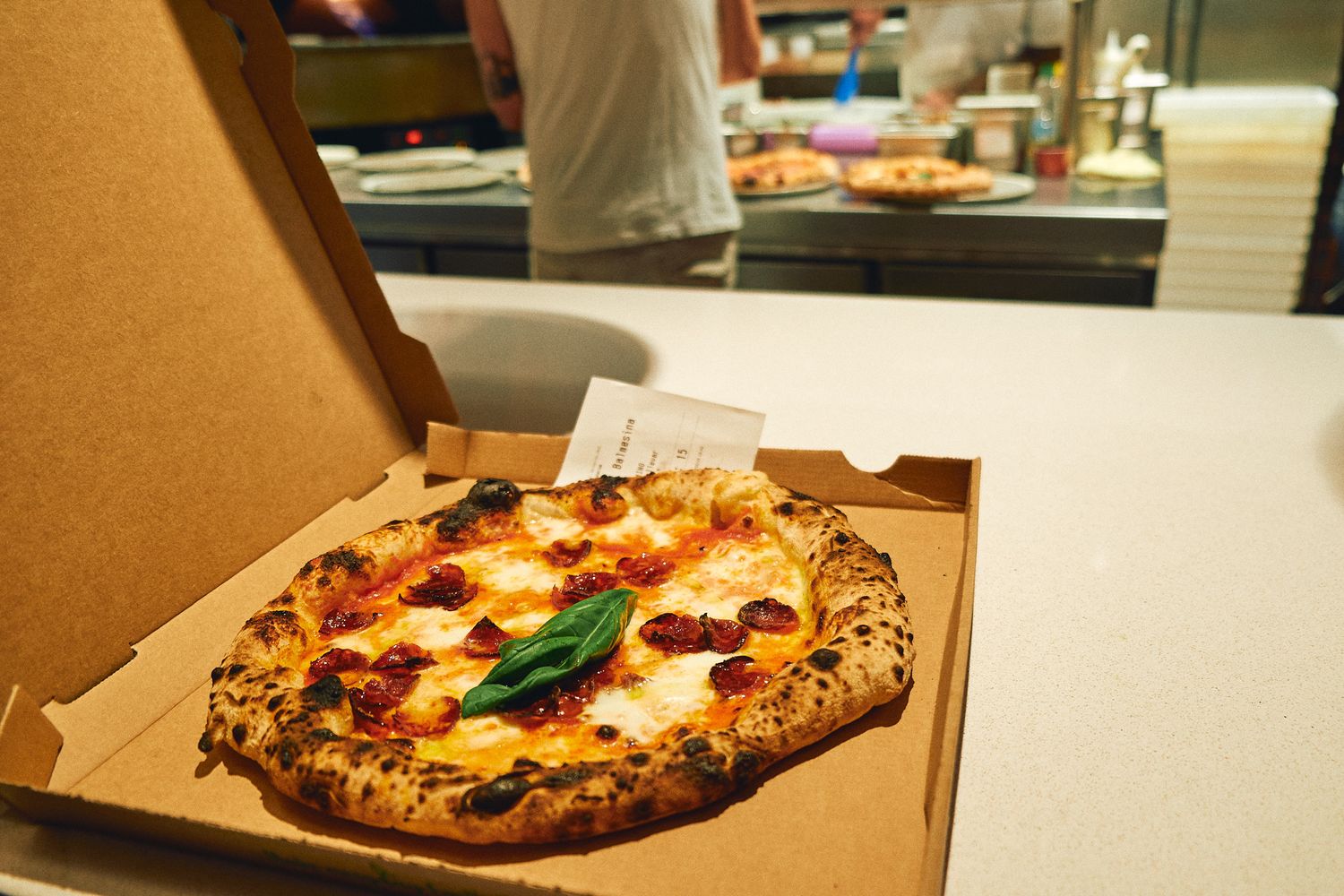 A freshly baked Margherita pizza with basil leaves in an open cardboard box on a white counter in a pizzeria.