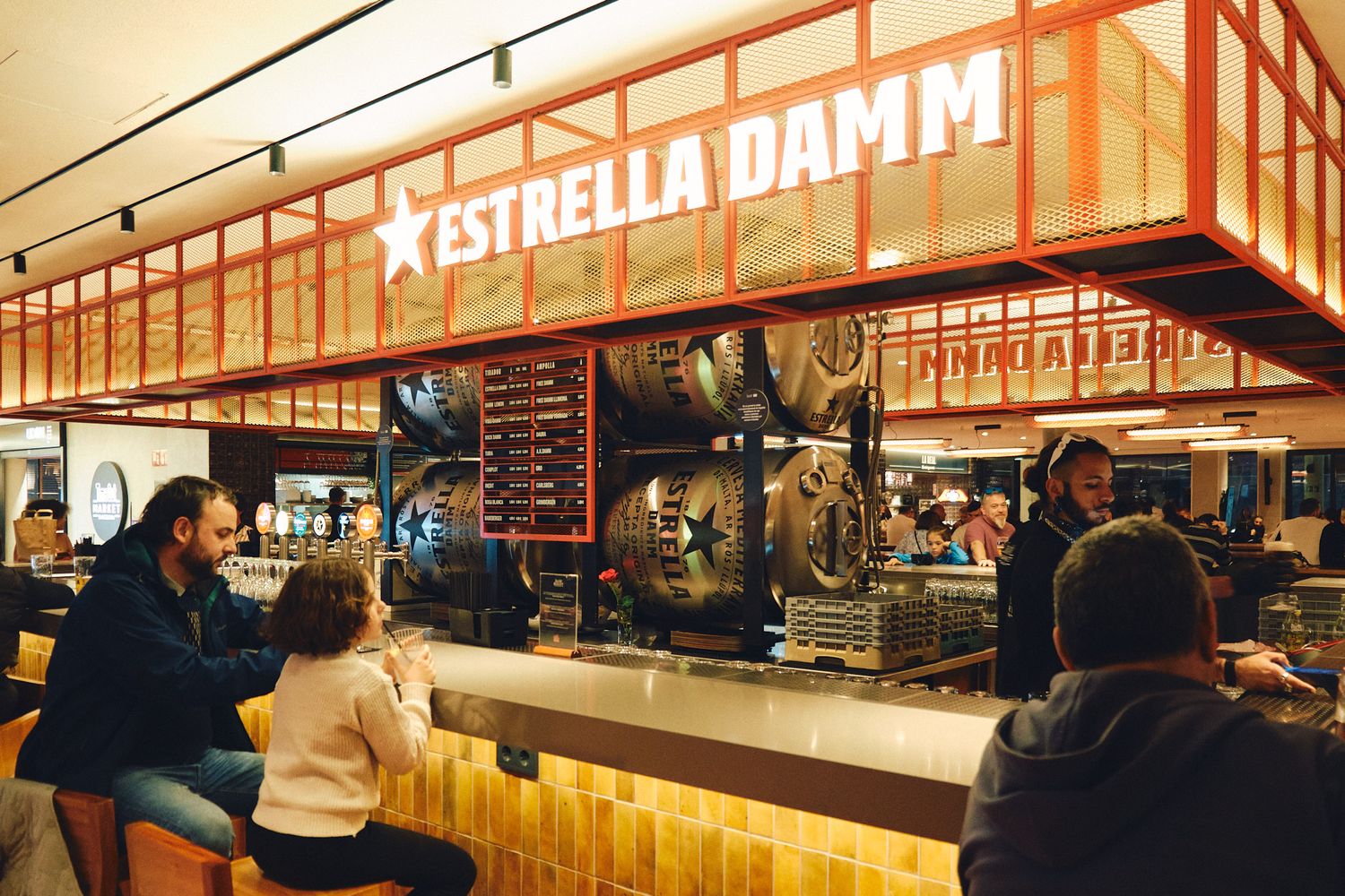 Estrella Damm bar with patrons seated at a wooden counter, featuring beer taps and industrial decor.