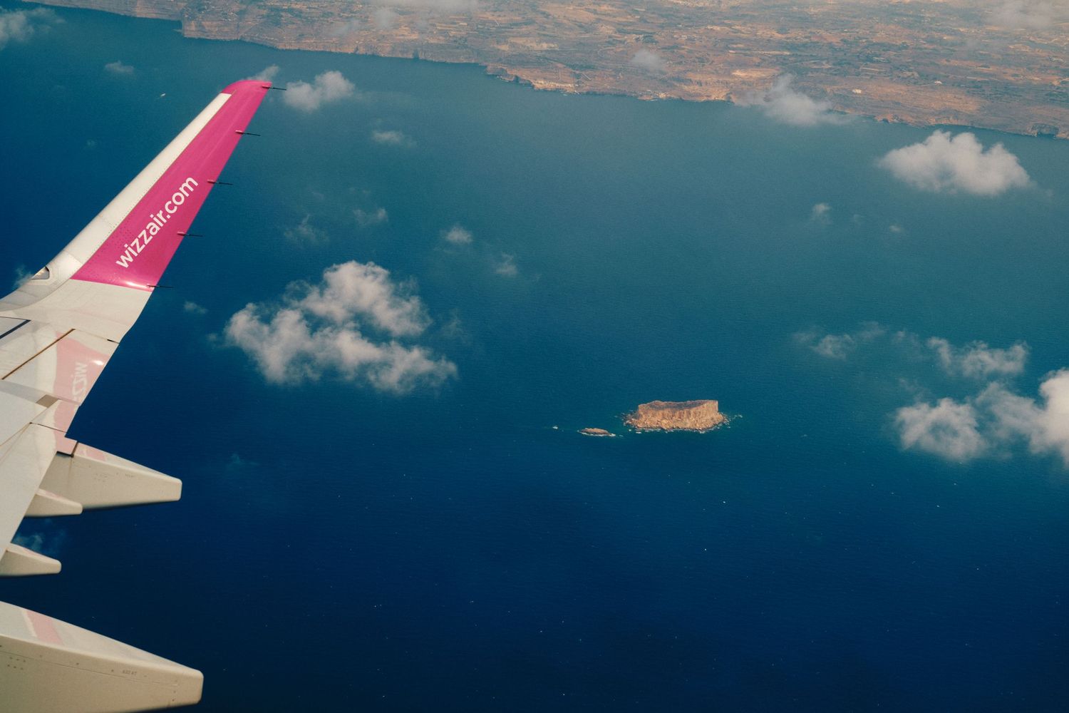 Pink airplane wing flying over blue ocean and small island.