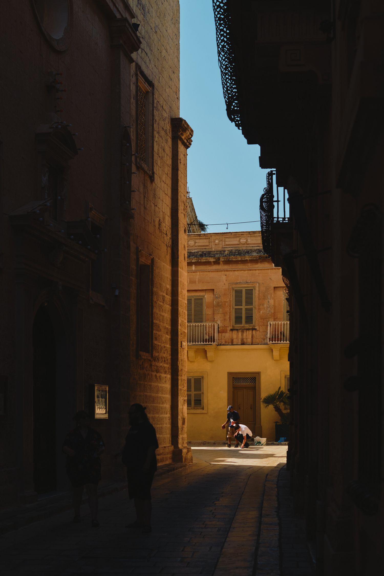 Narrow alley between historic stone buildings with sunlit courtyard at the end.