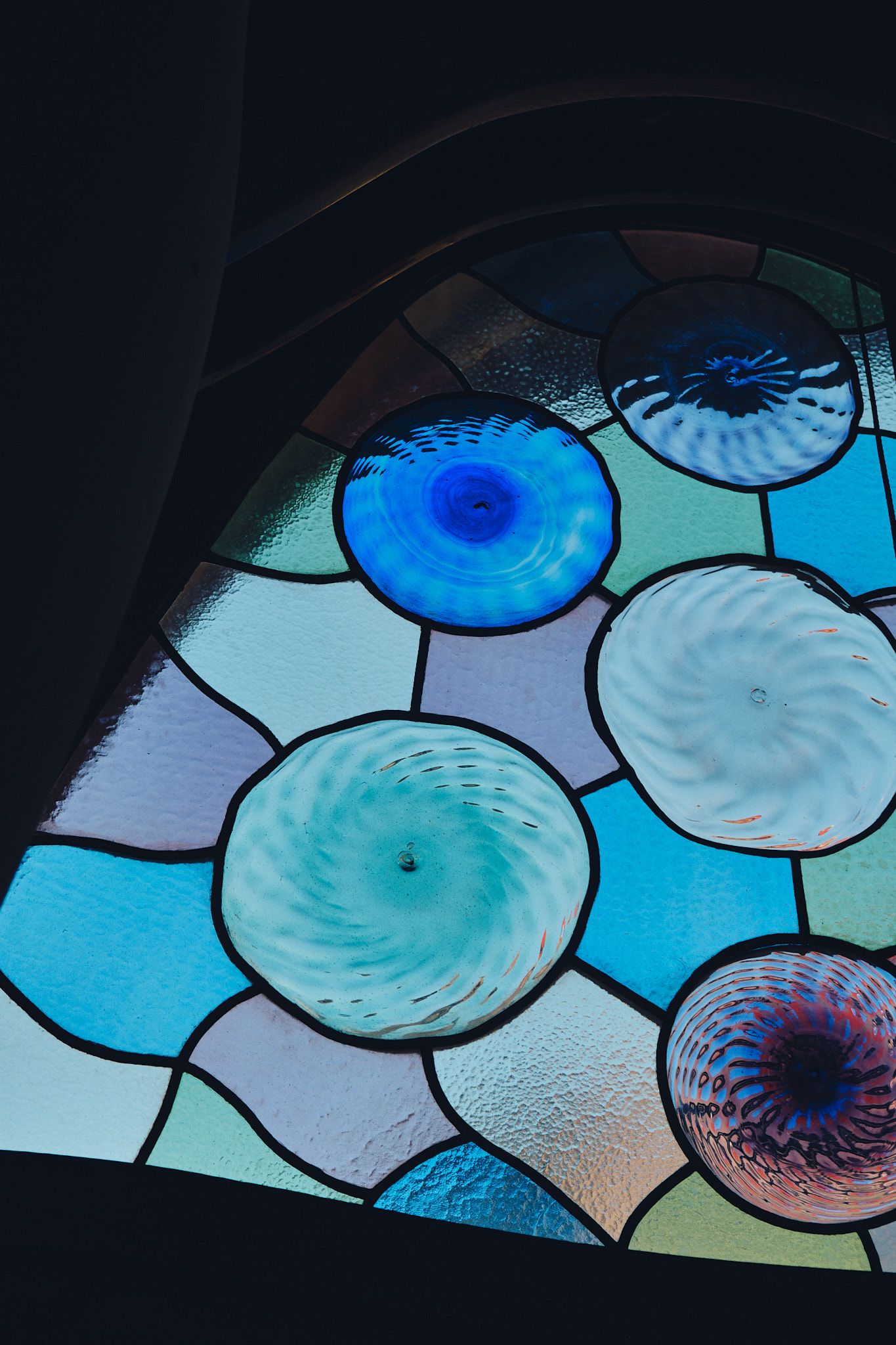 Stained glass window featuring circular swirled glass pieces in shades of blue, teal, and purple on a light blue background.