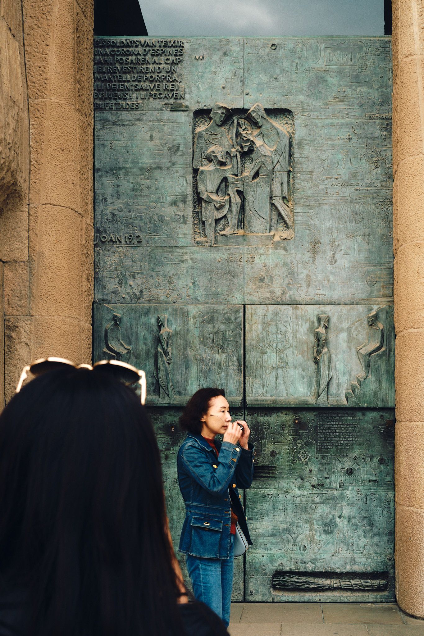 Woman in denim jacket photographing ornate bronze relief sculpture panels on a stone cathedral wall.