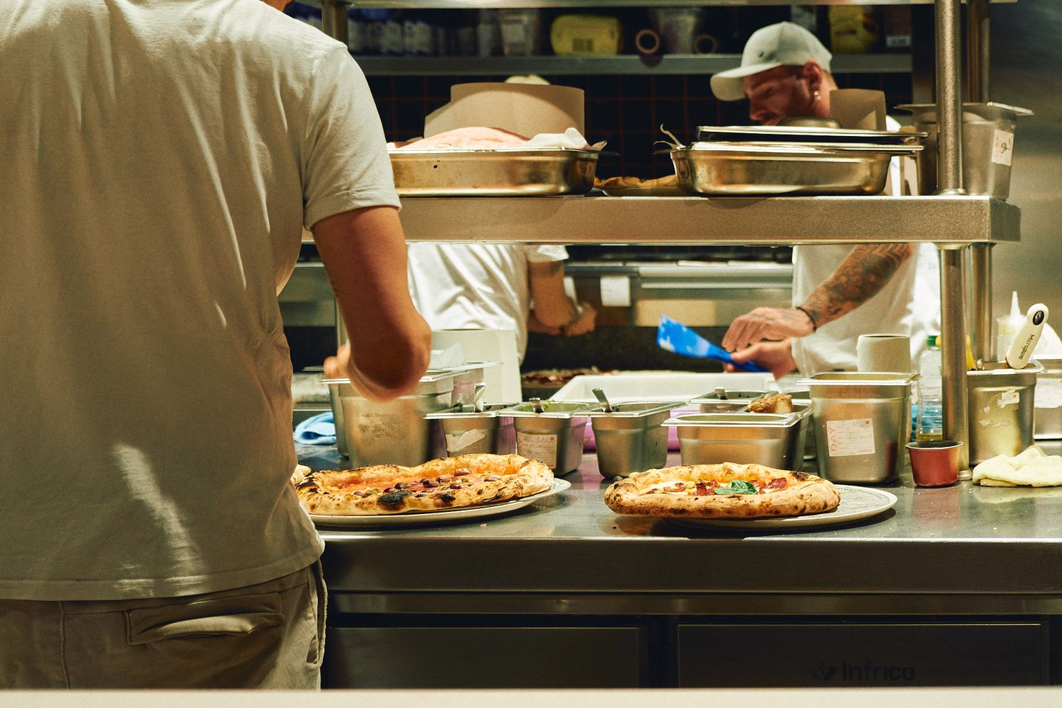 Two chefs preparing fresh pizzas in a busy restaurant kitchen with stainless steel counters and warm lighting.