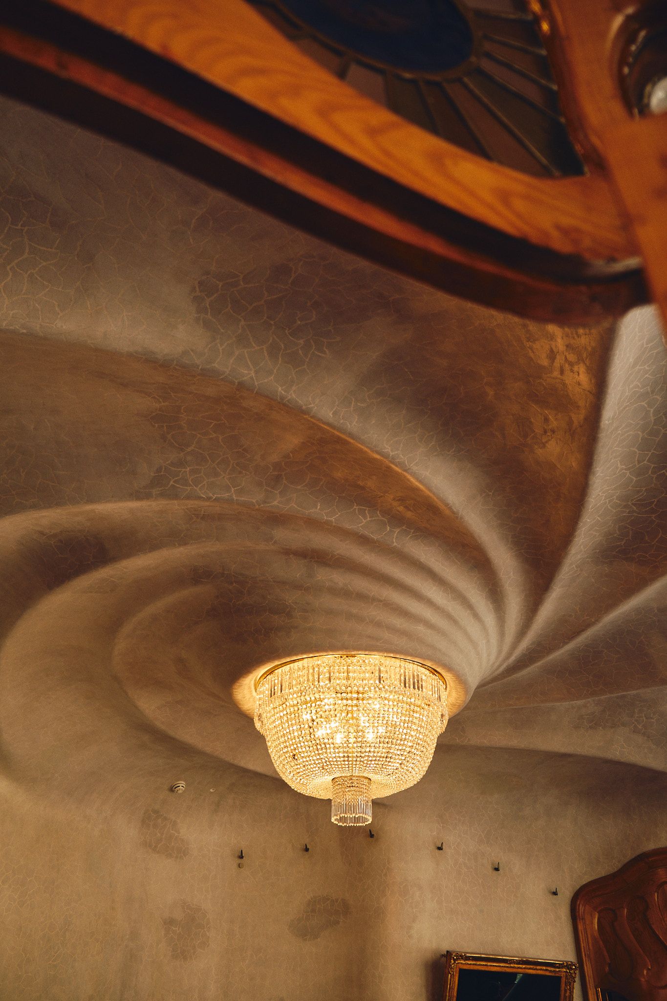 Crystal chandelier hanging from an ornate swirling plaster ceiling with warm golden light in an elegant historic interior.