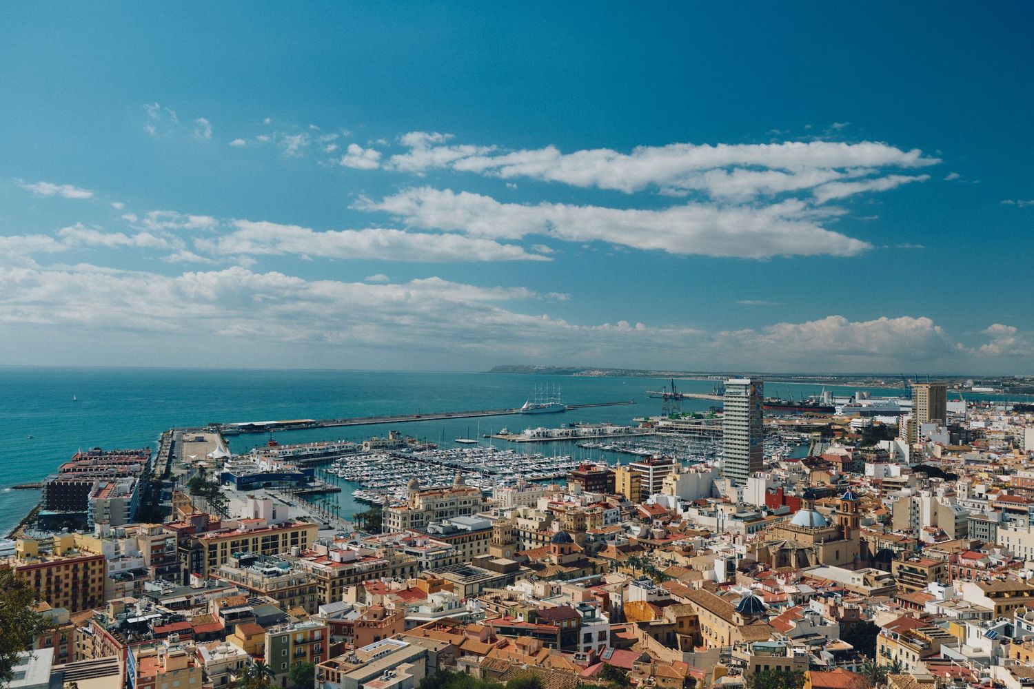 Aerial view of coastal city with modern port facilities and historic buildings against blue Mediterranean Sea.