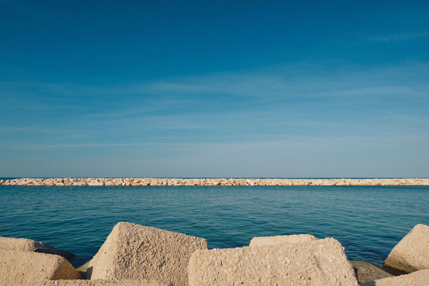 Concrete breakwater extends into calm blue sea under bright sky.