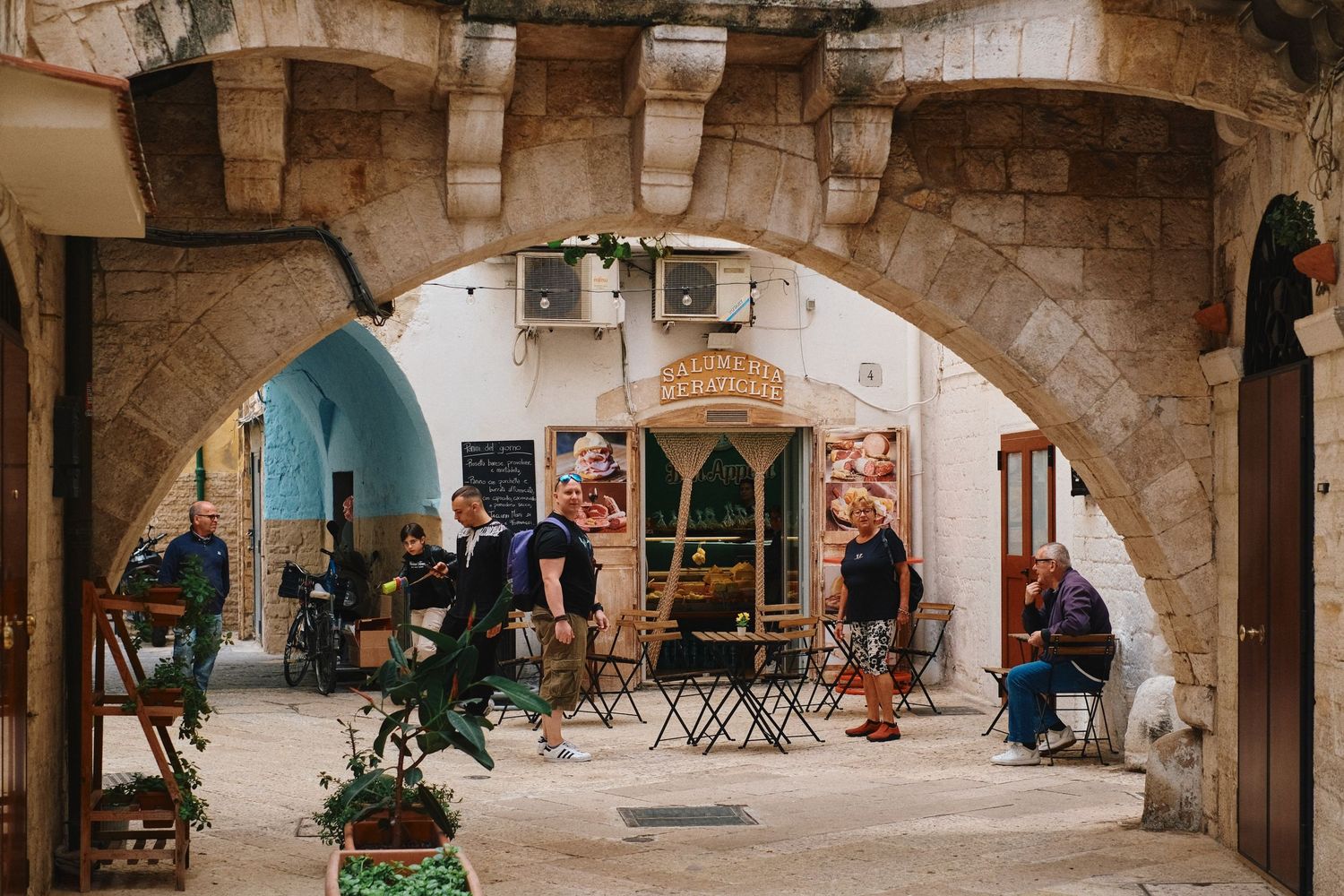 People relaxing at cafe tables under stone archway in historic European setting.