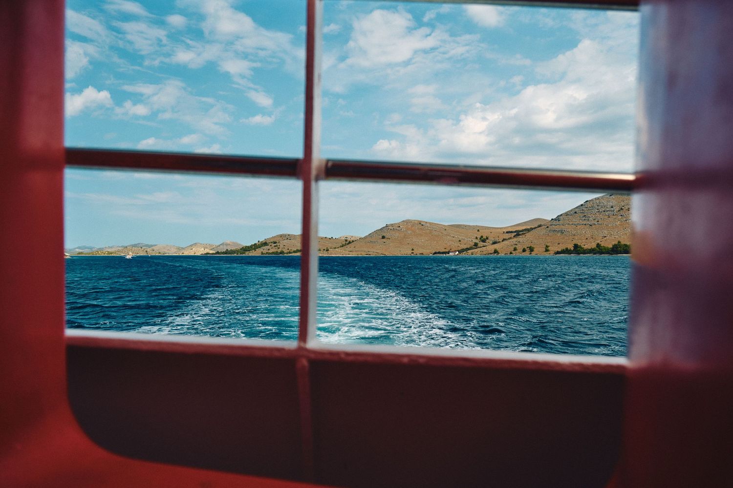 View of a Mediterranean coastline through a ship window showing blue water and rugged terrain.