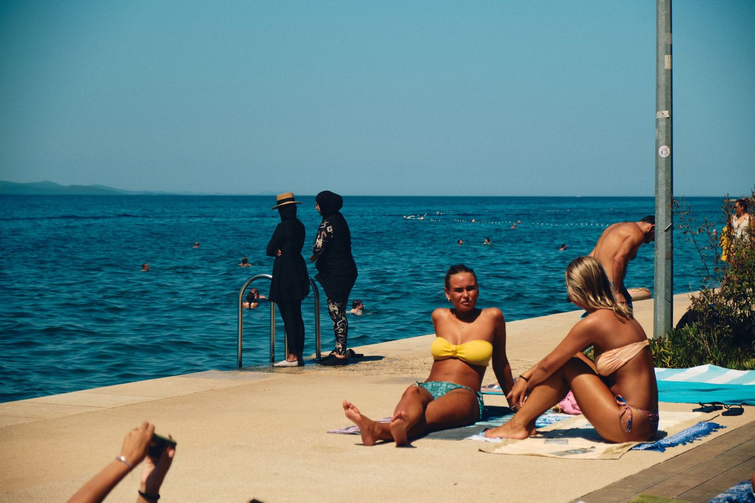 People relaxing on a sunny concrete pier overlooking turquoise Mediterranean waters.
