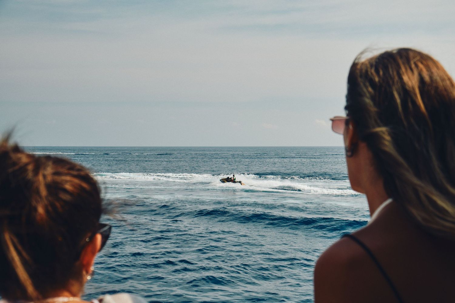 Two people watching a boat sail across blue Mediterranean waters from deck viewpoint.