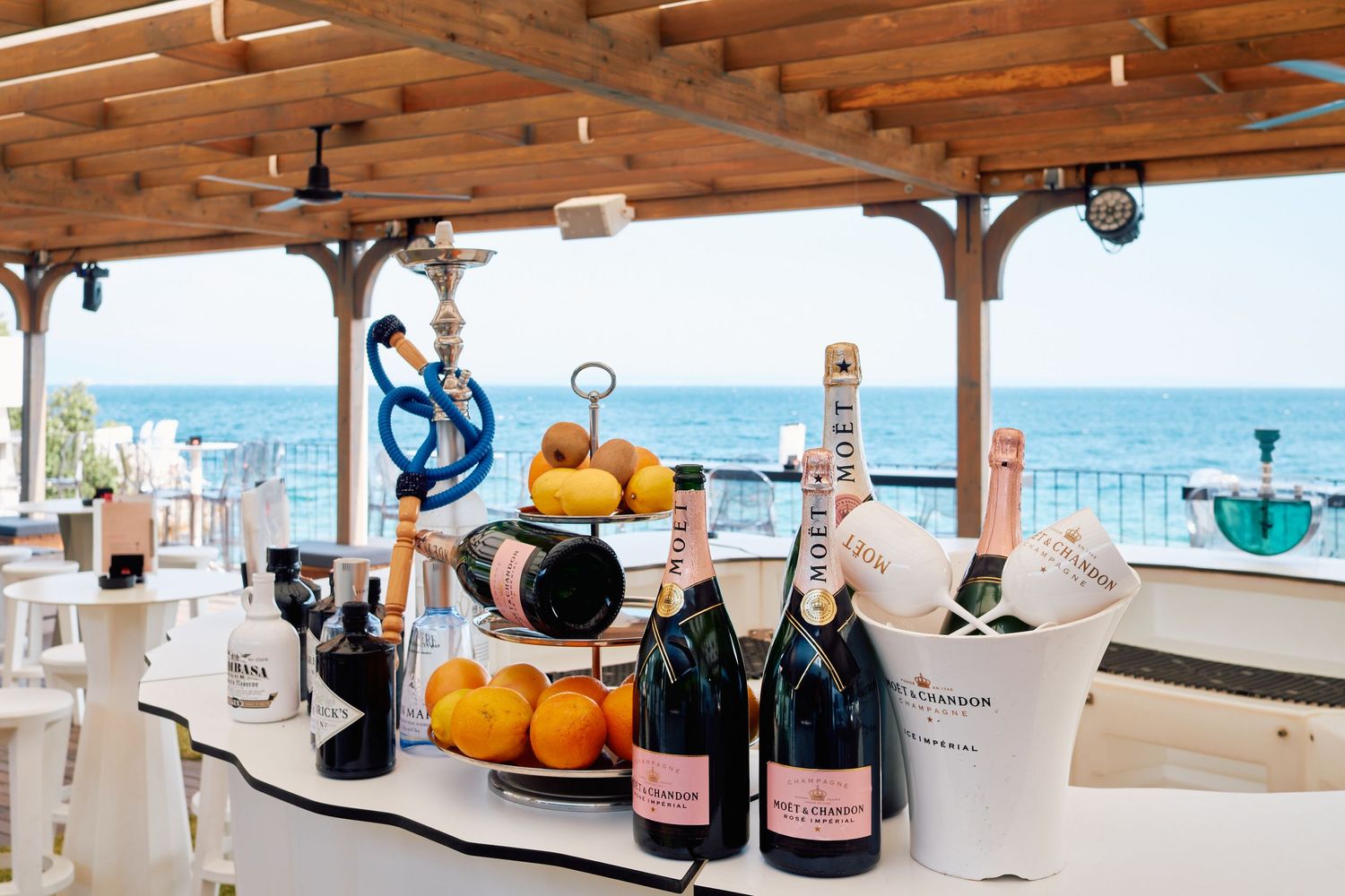 Seaside bar setup with champagne bottles and bar accessories under a wooden pergola.
