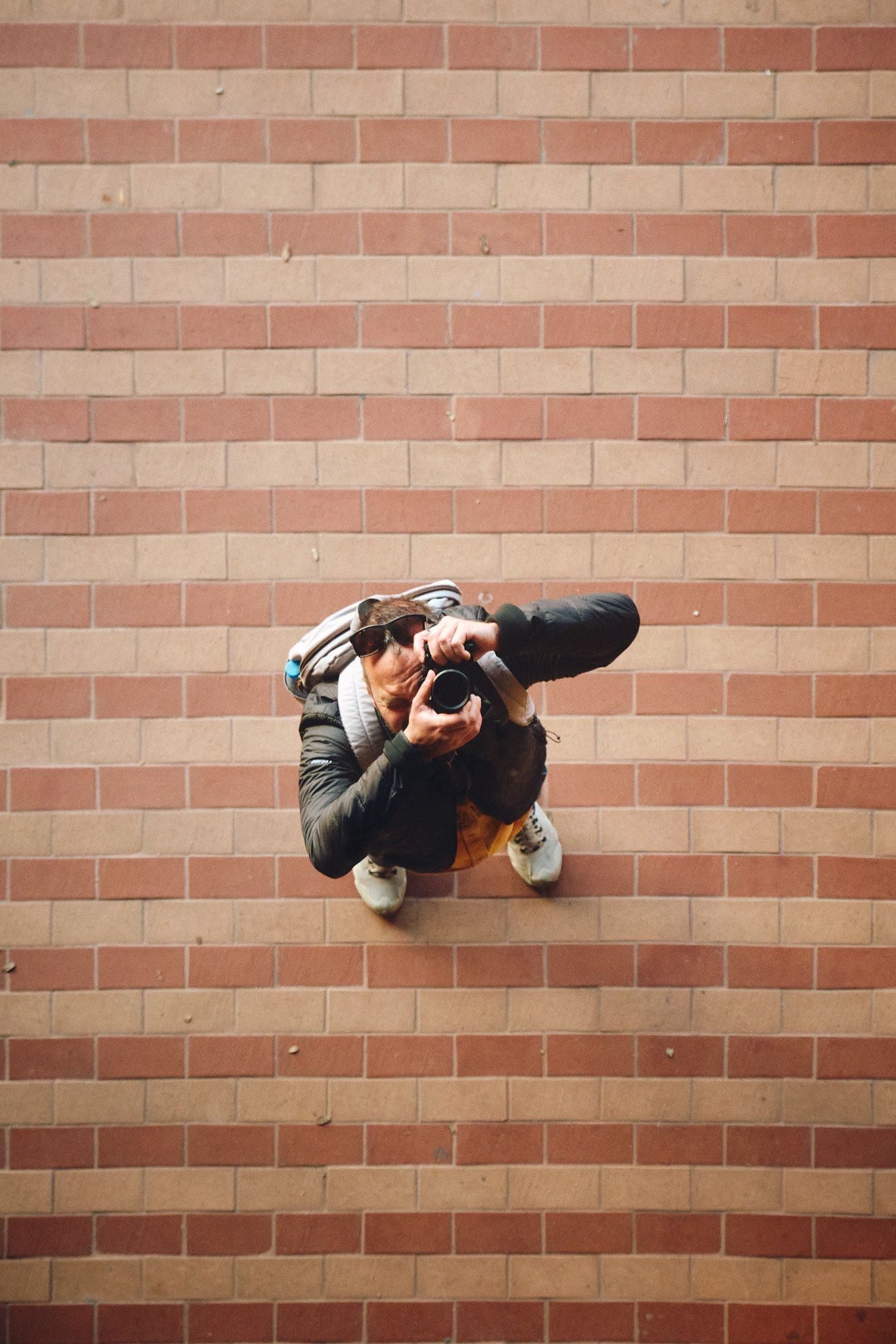 Aerial view of a photographer holding a camera, standing on a brick floor wearing a backpack and white sneakers.