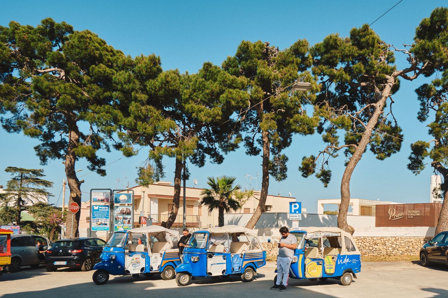 Blue tuk-tuk taxis parked under pine trees on a sunny street in a Mediterranean town.