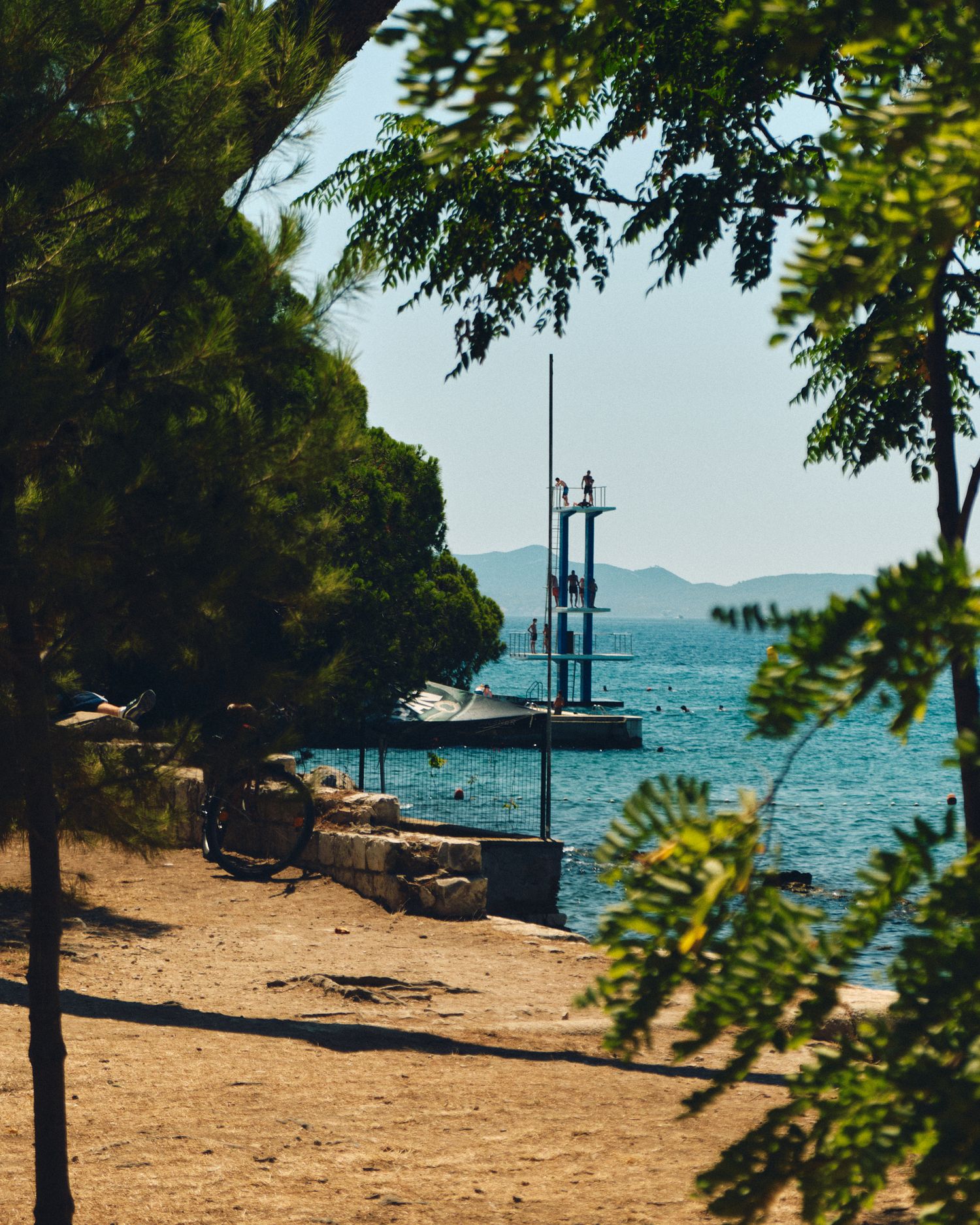 Scenic coastal path with boats and mountains visible through tree branches.