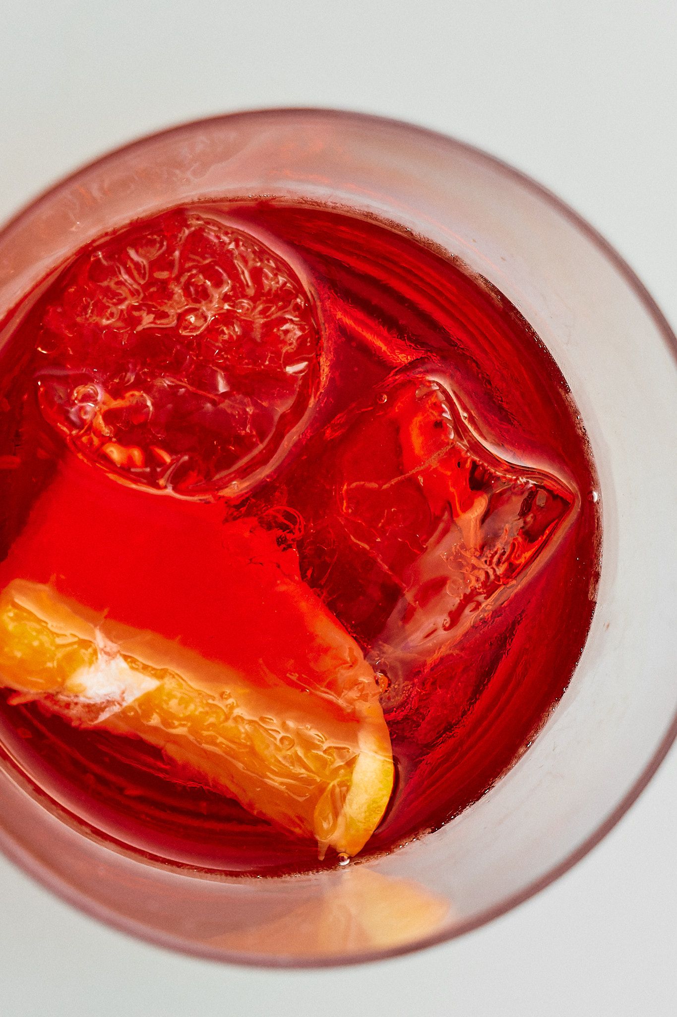 Overhead view of a vibrant red Negroni cocktail with ice cubes and an orange peel garnish in a glass.